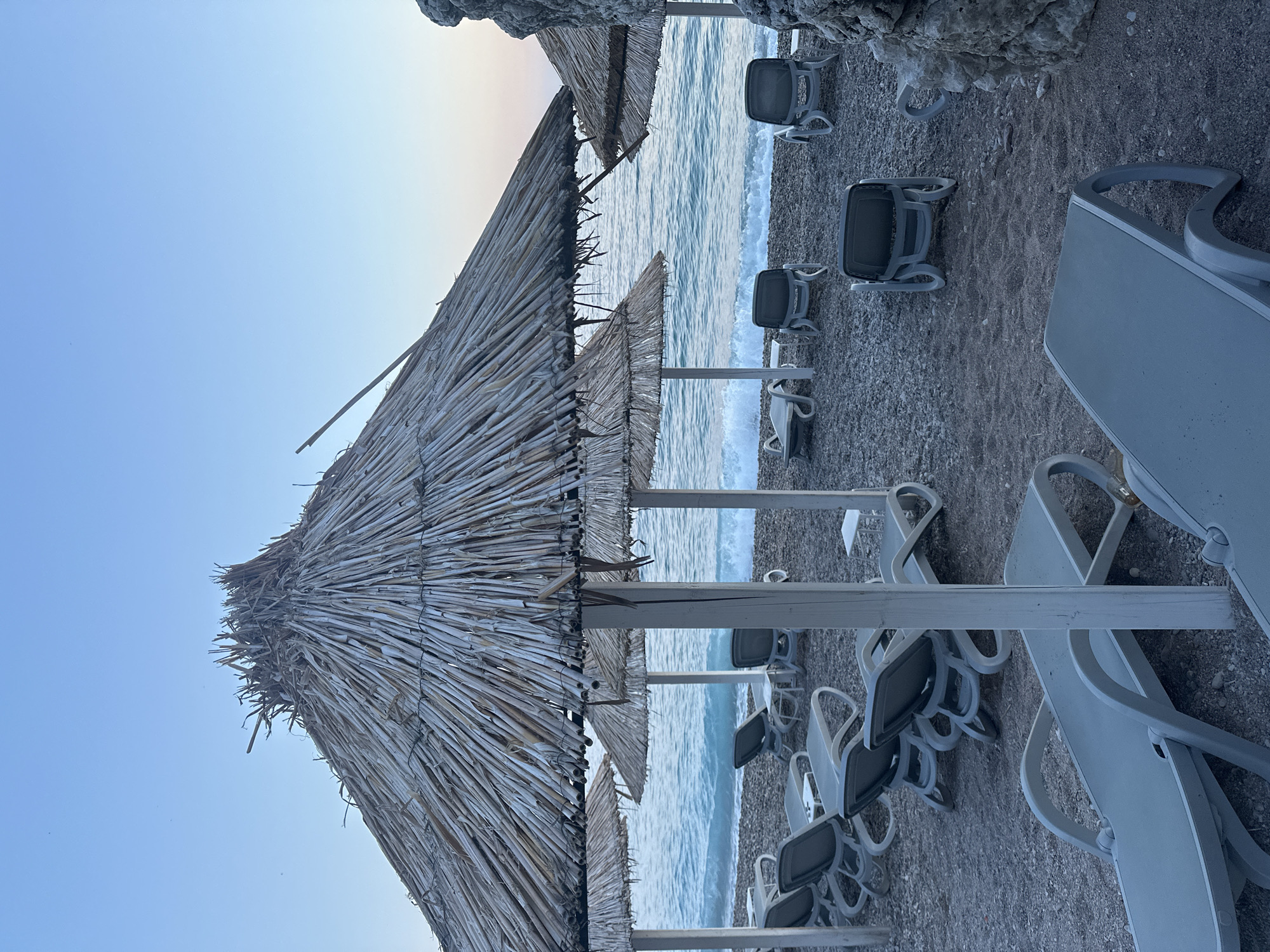 The image shows a beach scene in Mirište, Montenegro, likely during the early morning or late evening given the soft, diffused light and the clear, pale blue sky. The foreground is dominated by rows of empty, grey plastic sun loungers on a pebble beach. Several straw-thatched umbrellas provide shade over some of the loungers. The umbrellas are supported by white wooden poles. Beyond the loungers and umbrellas, the turquoise sea stretches to the horizon, with gentle waves lapping the shore. The water appears calm. The beach itself is covered in small, dark pebbles. To the right of the image, a large, grey rock formation juts out, partially obscuring a straw umbrella and the sea. The overall atmosphere is serene and unoccupied.