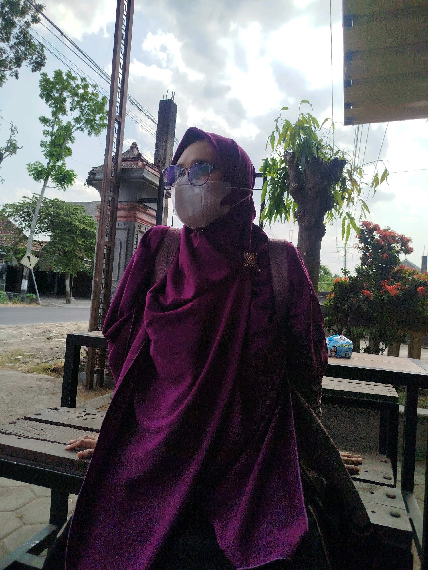 A medium shot captures a person wearing a purple hijab and a light-colored face mask, along with eyeglasses, posing outdoors in Karangwuni, Indonesia. The individual is seated at a rustic wooden table and bench set. The person's left hand rests on the table. Behind them, lush green trees and foliage create a natural backdrop, partially obscuring a building with traditional Indonesian architecture. The sky above is partly cloudy, with a mix of white clouds and blue sky visible. Power lines crisscross the scene, indicating an urban or semi-urban environment. The overall setting suggests a casual outdoor setting, possibly a cafe or a roadside rest area.