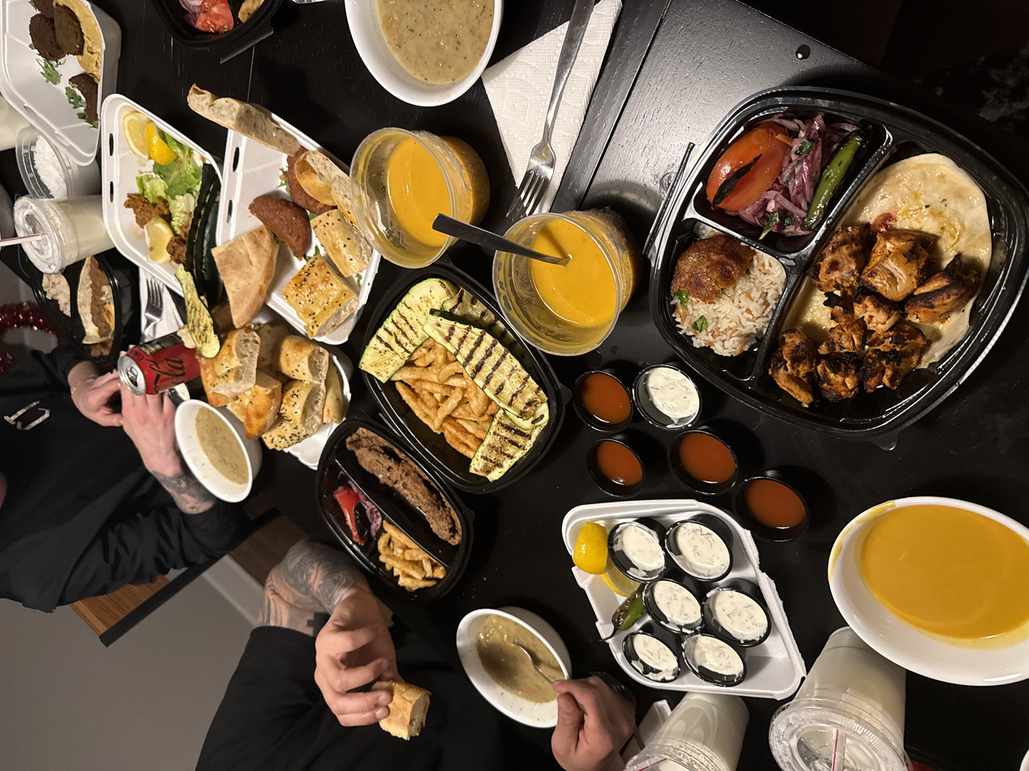 The image shows a table covered with numerous containers of food, most of which appear to be Middle Eastern cuisine. Visible items include various dips, bread, grilled vegetables, soup, rice, chicken, and salads. Two people are partially visible, with their hands and arms present in the foreground, suggesting they are eating or preparing to eat the food. One person is holding a piece of bread. A Coca-Cola can is also visible. Based on the variety and style of food, it could be from a Middle Eastern restaurant or a catered meal. The exact location in Chicago cannot be determined from the image alone.