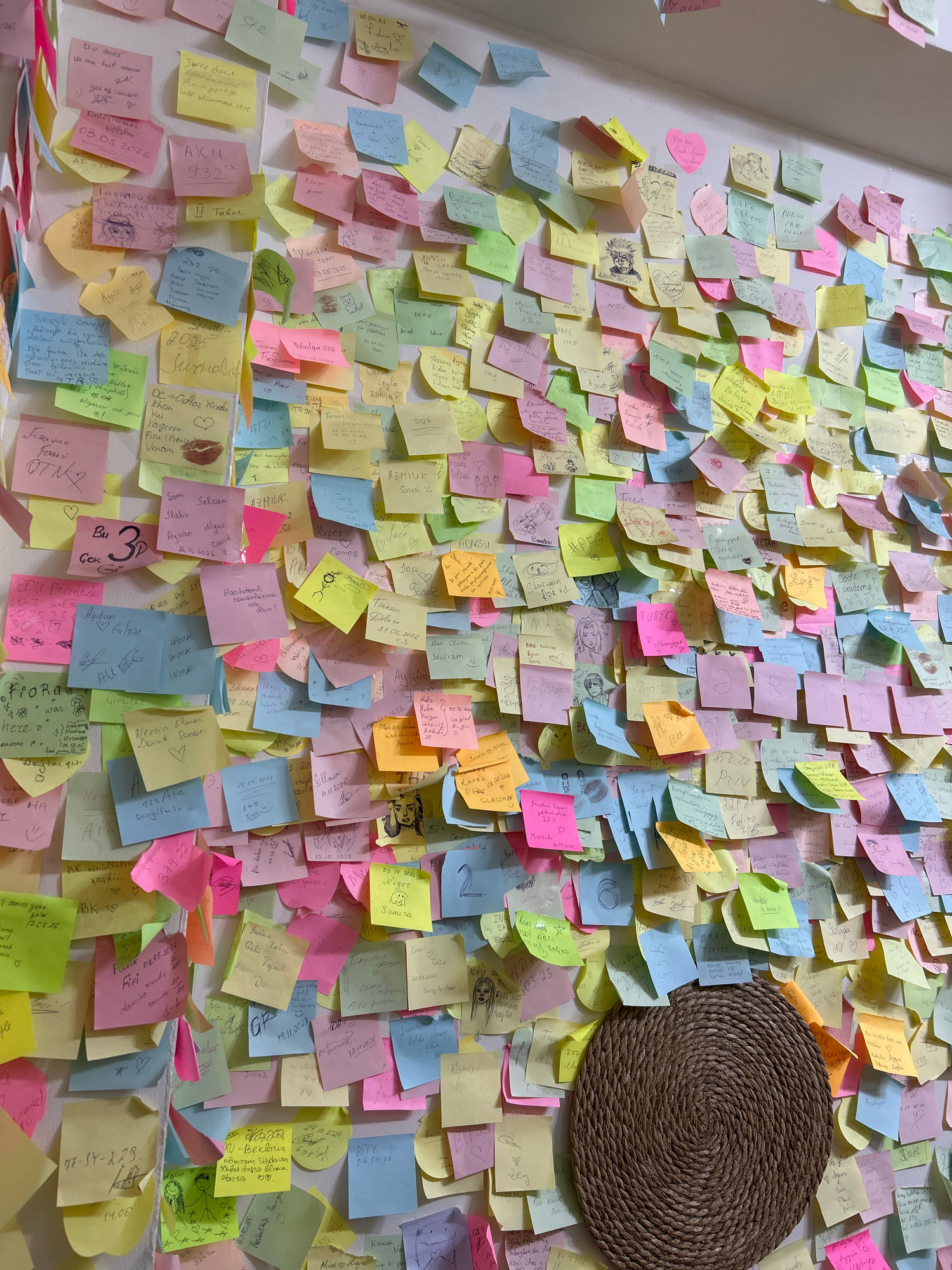 The image depicts a wall densely covered with numerous colorful sticky notes in various sizes and orientations. The notes are handwritten and some contain drawings. The overall impression is that of a highly personalized and interactive display, likely a message board or a space for shared thoughts and memories. The text on the notes suggests a mix of languages, including Azerbaijani and possibly Turkish, with some dates visible.
The sticky notes are affixed to a plain, light-colored wall, which appears to be indoors. The lighting is diffuse, suggesting ambient room light, and there are no strong shadows. The composition is filled with the sticky notes, creating a textured and visually busy surface.
In the lower right quadrant of the image, a circular, woven mat made of natural fibers is partially visible, adding a contrasting organic texture to the scene. The mat is brown and appears to be a decorative element.
The arrangement of the notes suggests a dynamic and ongoing process of communication, with some notes overlapping others. The sheer volume of notes implies a significant amount of activity and participation over time.
While the image does not contain explicit visual markers identifying the specific location in Baku, Azerbaijan, the presence of Azerbaijani language on some of the notes strongly suggests this as the geographical context. This kind of display is commonly found in public spaces or community hubs within cities, serving as a platform for shared experiences and connections.