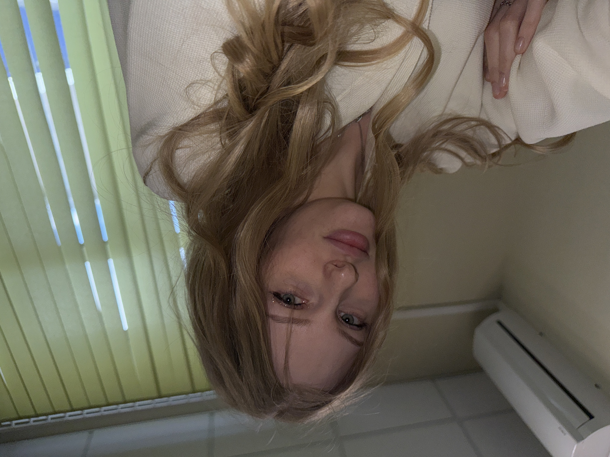 A woman with long, blonde, wavy hair is photographed from an inverted perspective. She is wearing a white knitted garment, possibly a sweater or robe. Her face is visible, featuring fair skin, light eyes, and pink lips. Light reflects off her skin and hair. A window with light green blinds is visible in the background. Also in the background is a white air conditioner unit. The setting appears to be indoors, possibly in a residential or hotel room. The location, Perm, Russia, is not determinable from the image.