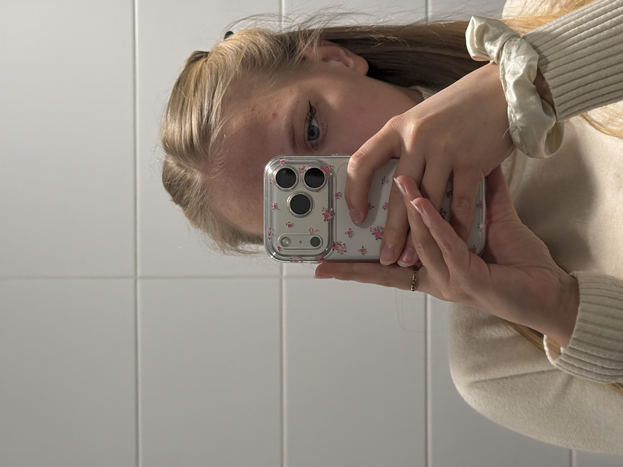 A woman is taking a selfie in front of a white tiled wall. She is holding a phone with a clear floral case. She has blonde hair and is wearing a light-colored sweater with a ruffled cuff. A gold ring is visible on her finger. There is no information to suggest the location is in Izhevsk, Russia.