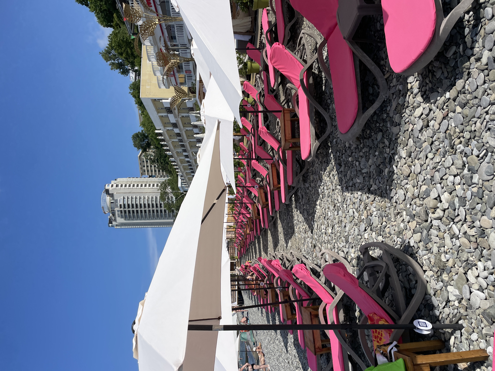 The image depicts a beach scene in Sochi, Russia. Numerous empty, pink-cushioned lounge chairs are neatly arranged on a pebble beach, shaded by large white umbrellas. In the background, a multi-story hotel with balconies and trees lines the shore, with a taller, modern building further back. The sky is a clear, bright blue, suggesting a sunny day, likely in the late morning or early afternoon given the strong shadows. A few people are visible on the beach, some lounging or walking, their presence adding a sense of casual leisure to the setting. The overall atmosphere is one of a typical resort beach, prepared for visitors seeking relaxation under the sun. There is a QR code visible on a small stand.