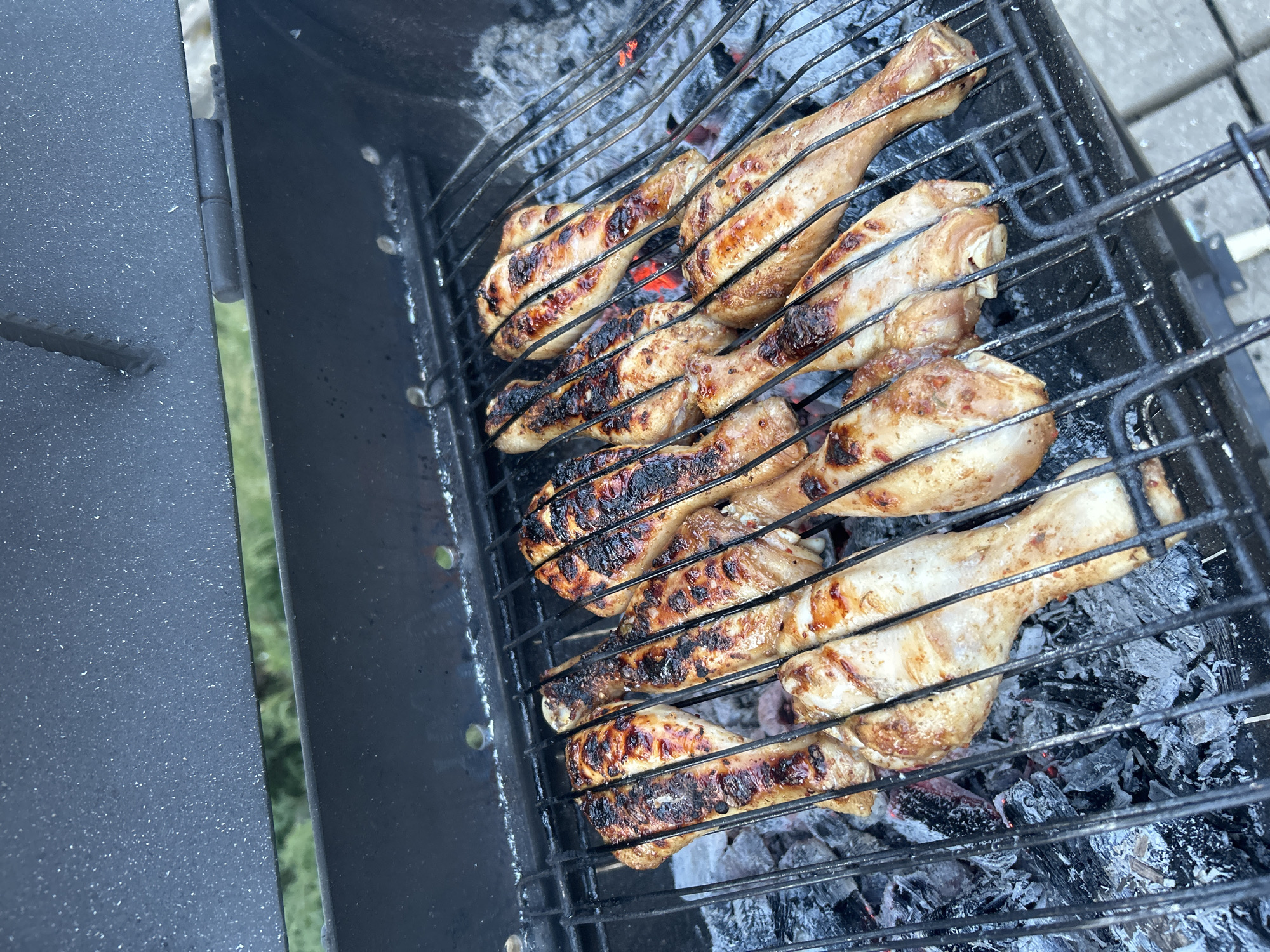 The image shows chicken drumsticks being grilled on a charcoal grill. Some of the drumsticks are charred, while others are less cooked. The grill is black and sits over a bed of gray ash and embers. The location is unknown.