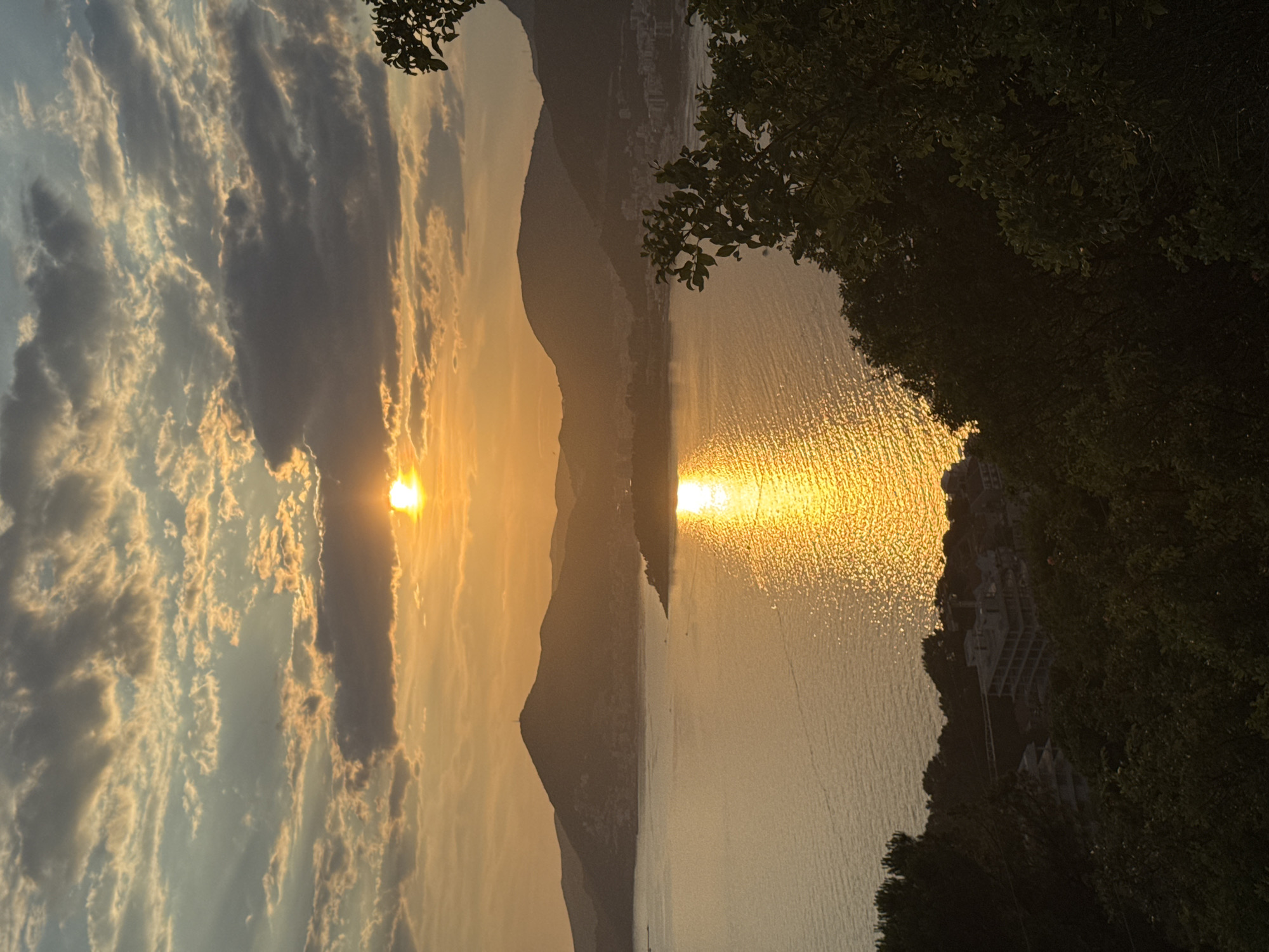 The image captures a scenic sunset over a body of water, likely a bay or gulf, with mountains in the background. The sun, a bright orb of light, is partially obscured by clouds, casting a golden-orange glow across the sky and reflecting a shimmering path on the water's surface. The water appears calm with gentle ripples, creating a textured reflection of the sun.
The foreground is dominated by lush green foliage, obscuring part of the view. In the middle ground, a coastline is visible with hilly terrain leading down to the water. Buildings, some appearing to be under construction with visible concrete structures, are scattered along the coastline, interspersed with trees. The mountains in the background are silhouetted against the vibrant sky, with their peaks softly rounded and some appearing to have a slightly lighter hue due to atmospheric haze. The clouds are a mix of darker, denser formations and lighter, wispy ones, tinged with the warm colors of the sunset.
There are no people or specific actions depicted in the scene. The overall impression is one of natural beauty and tranquility at dusk. The specific location within "Unknown, Unknown" cannot be determined from the visual information provided.