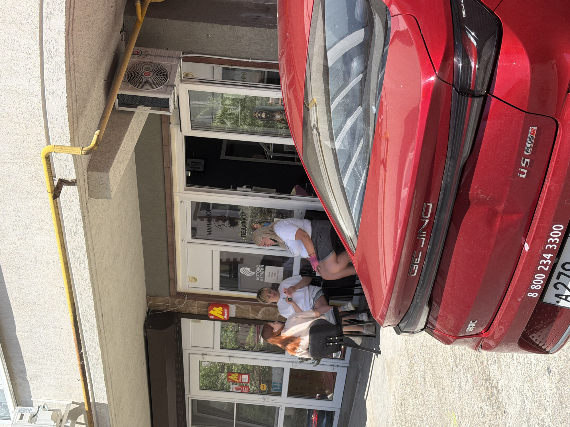 The image shows a street scene in Sochi, Russia, with a red car parked in the foreground, obscuring the lower half of a building. The building appears to be a commercial establishment, possibly a salon or shop, with large glass doors and windows. Three women are seated outside the establishment, seemingly engaged in conversation. The woman on the left has red hair and is wearing a light-colored top. The woman in the middle, with blonde hair, is wearing a white t-shirt and denim shorts and is looking at her phone. The woman on the right has blonde hair, is wearing a white t-shirt and a grey skirt, and is wearing pink gloves. A yellow pipe runs horizontally along the building's facade, and an air conditioning unit is mounted on the wall. The ground appears to be paved with concrete. The car in the foreground is a red sedan with a "BEIJING" logo visible on its rear. There is also a phone number "8 800 234 3300" and "U5 PLUS" text on the car.