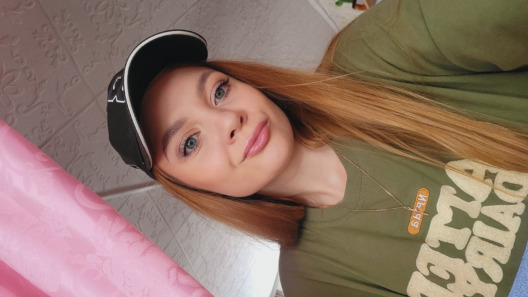 A young woman is posing for a selfie. She is wearing a green t-shirt with large, stylized lettering. A black baseball cap with white stitching is on her head. She has light skin, blue eyes, and reddish-blonde hair. She is wearing light makeup, including lip gloss. A thin gold necklace is visible around her neck. A pink curtain hangs to the left. The background behind her includes a textured, white wall. Зипуново, Russia is not discernable in the image.