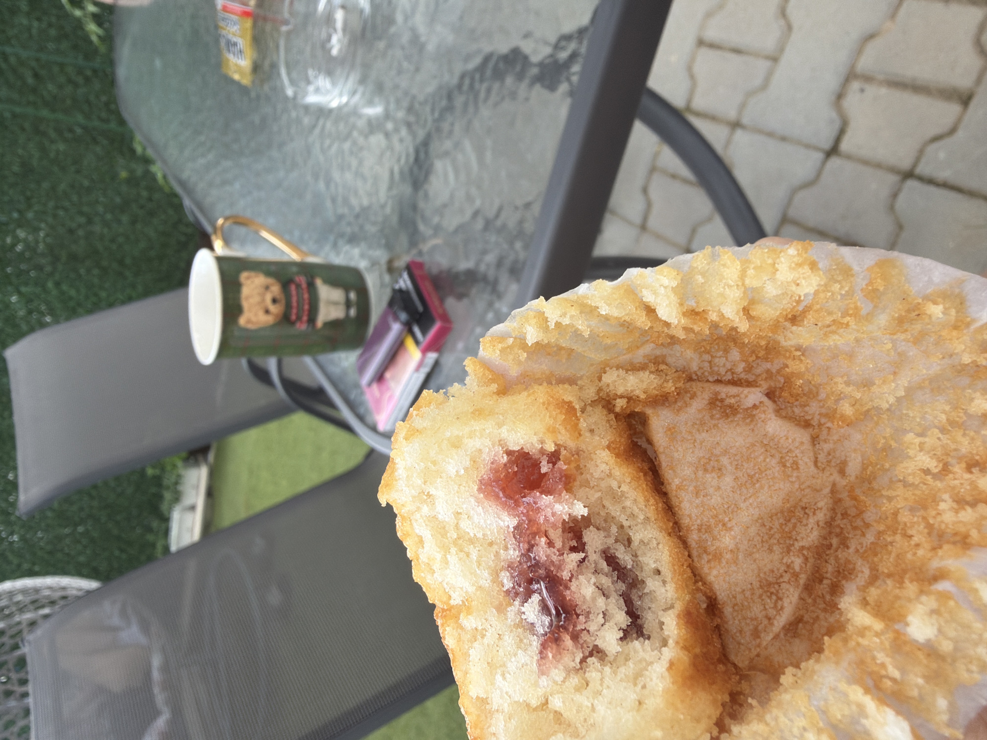 This media file captures a close-up view of a partially eaten cupcake with jam filling, held in a person's hand. The cupcake is placed on a paper liner, revealing its golden-brown, slightly crumbly texture and the vibrant red jam. In the background, a glass-top table is visible, with a mug featuring a teddy bear design and a pink and silver lighter resting on it. The setting appears to be outdoors, possibly on a patio or garden, with glimpses of green artificial turf and grey outdoor chairs. The lighting suggests it's daytime. There is no visible text on the cupcake or its liner. The context provided indicates the location is Sofia, Bulgaria.