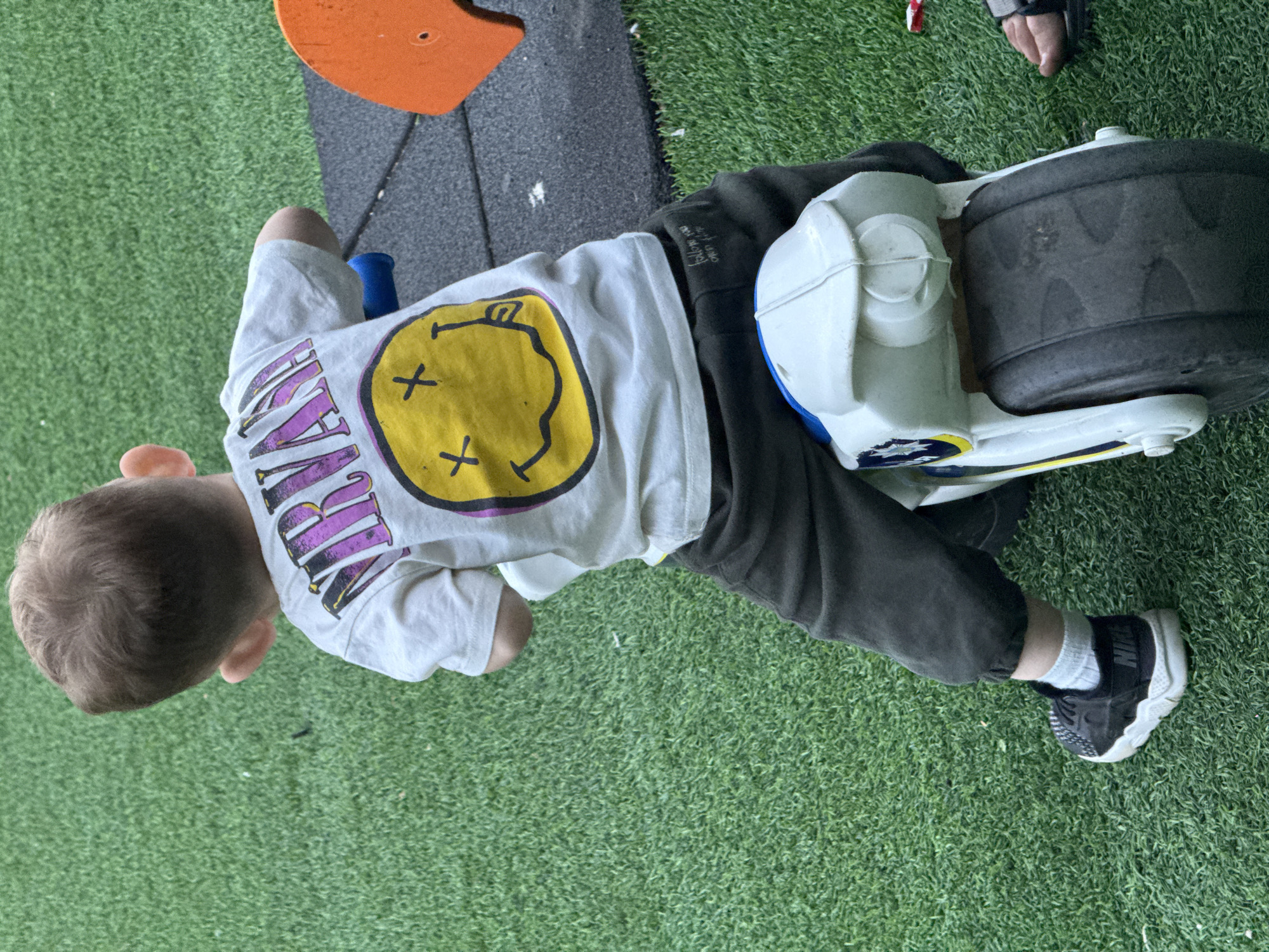 A young child, viewed from the rear, is seated on a white and blue toy motorcycle on an artificial turf surface. The child wears a white t-shirt with a "NIRVANA" logo and a smiley face graphic, dark pants, and black athletic shoes with white soles and light gray socks. The toy motorcycle has a large black rear tire. In the background, there is a black rubber mat and an orange playground structure. The overall setting appears to be an outdoor playground or recreational area.