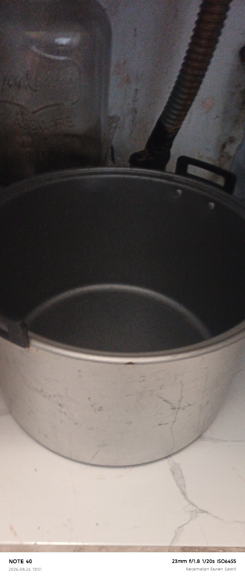 The image is a close-up shot of a large, silver pot with two dark handles. The pot appears to be made of metal and shows some signs of wear, with visible scratches and marks on its exterior. The interior of the pot is dark and appears to be the cooking surface. In the background, out of focus, is a large glass jar and a corrugated pipe. The lighting is dim, suggesting an indoor setting. At the bottom of the image, text reads "NOTE 40 2026.08.26 13:51" and "23mm f/1.8 1/20s ISO6455 Kecamatan Duren Sawit." This text indicates the photo was taken with a "Note 40" device, the date and time, camera settings, and the location as "Kecamatan Duren Sawit," which is a district in East Jakarta, Indonesia.