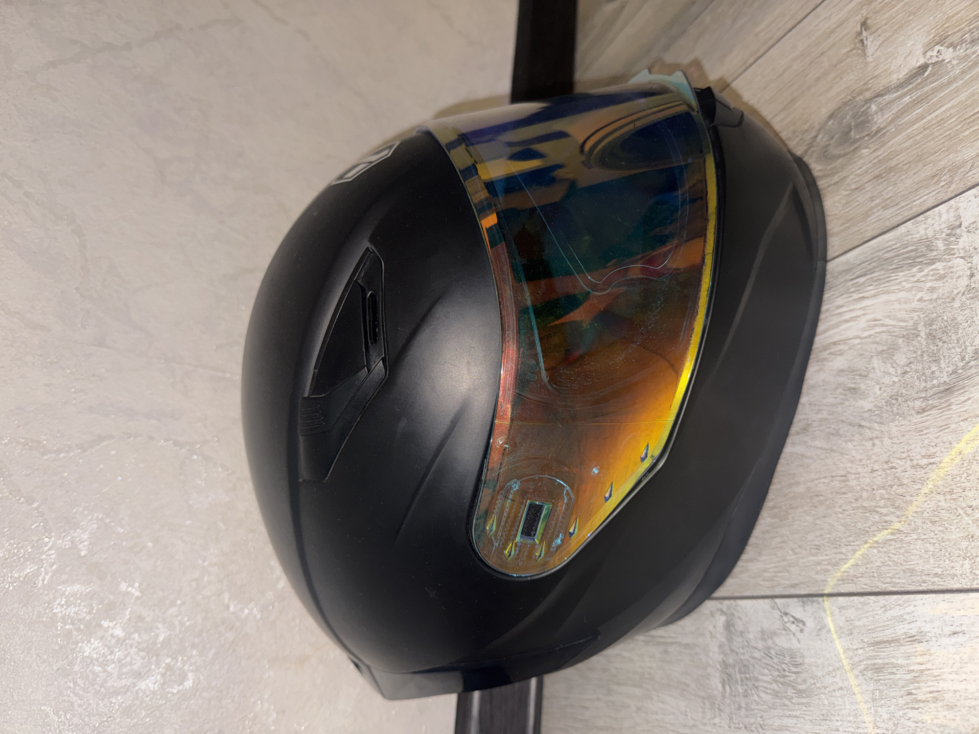 A black motorcycle helmet with a multicolored iridescent visor is placed on a light-colored wooden floor in front of a textured off-white wall. The visor reflects the surrounding environment with hues of yellow, orange, green, and blue. The helmet appears to be resting on its base. The scene is lit, casting faint shadows.