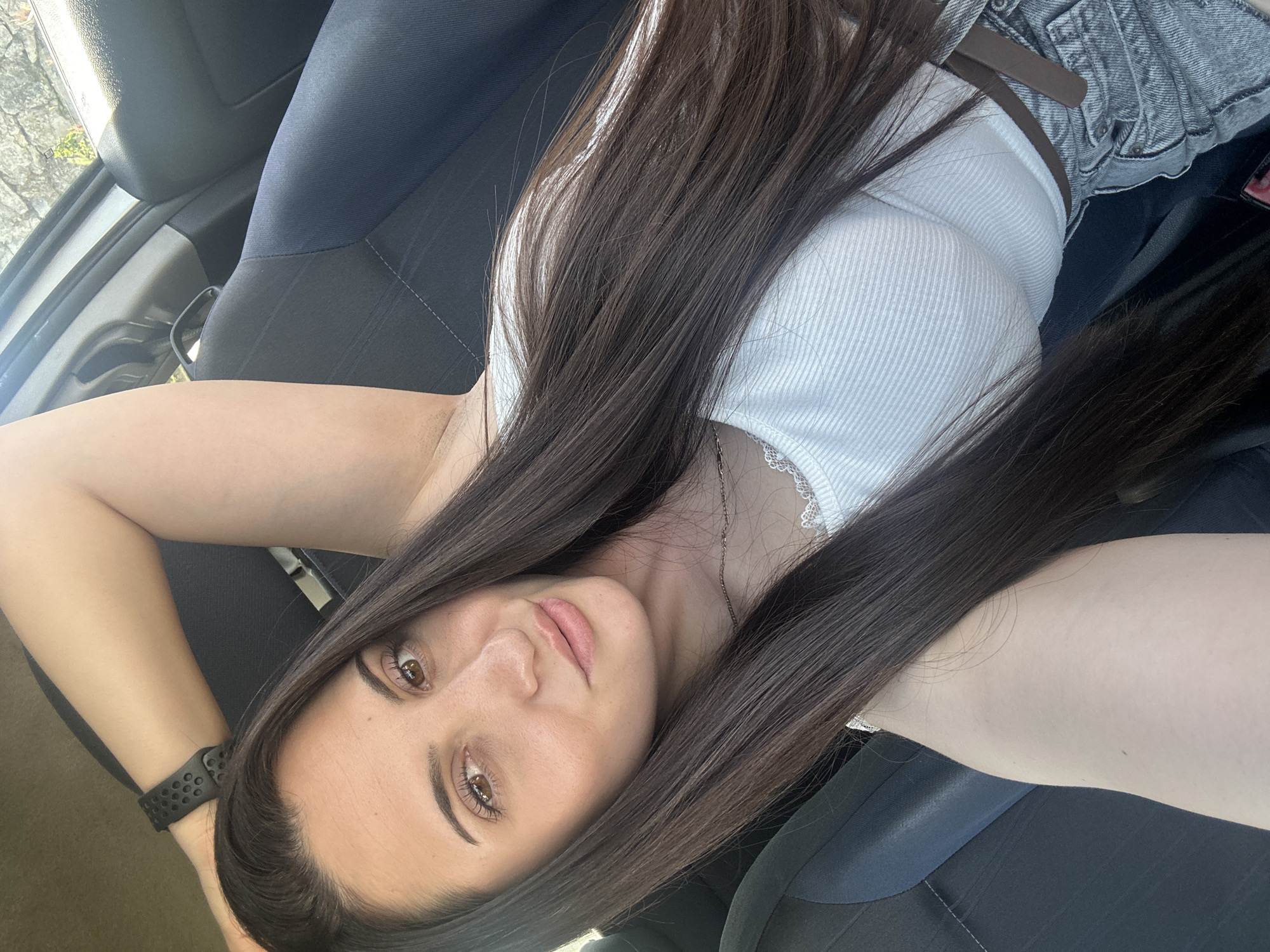 A young woman with long dark brown hair, wearing a white ribbed tank top and denim jacket, is lying in the front seat of a car. She is wearing a black smart watch on her left wrist. She is looking at the camera. The car appears to be parked, but the specific location in Beliakovets, Bulgaria is indiscernible.