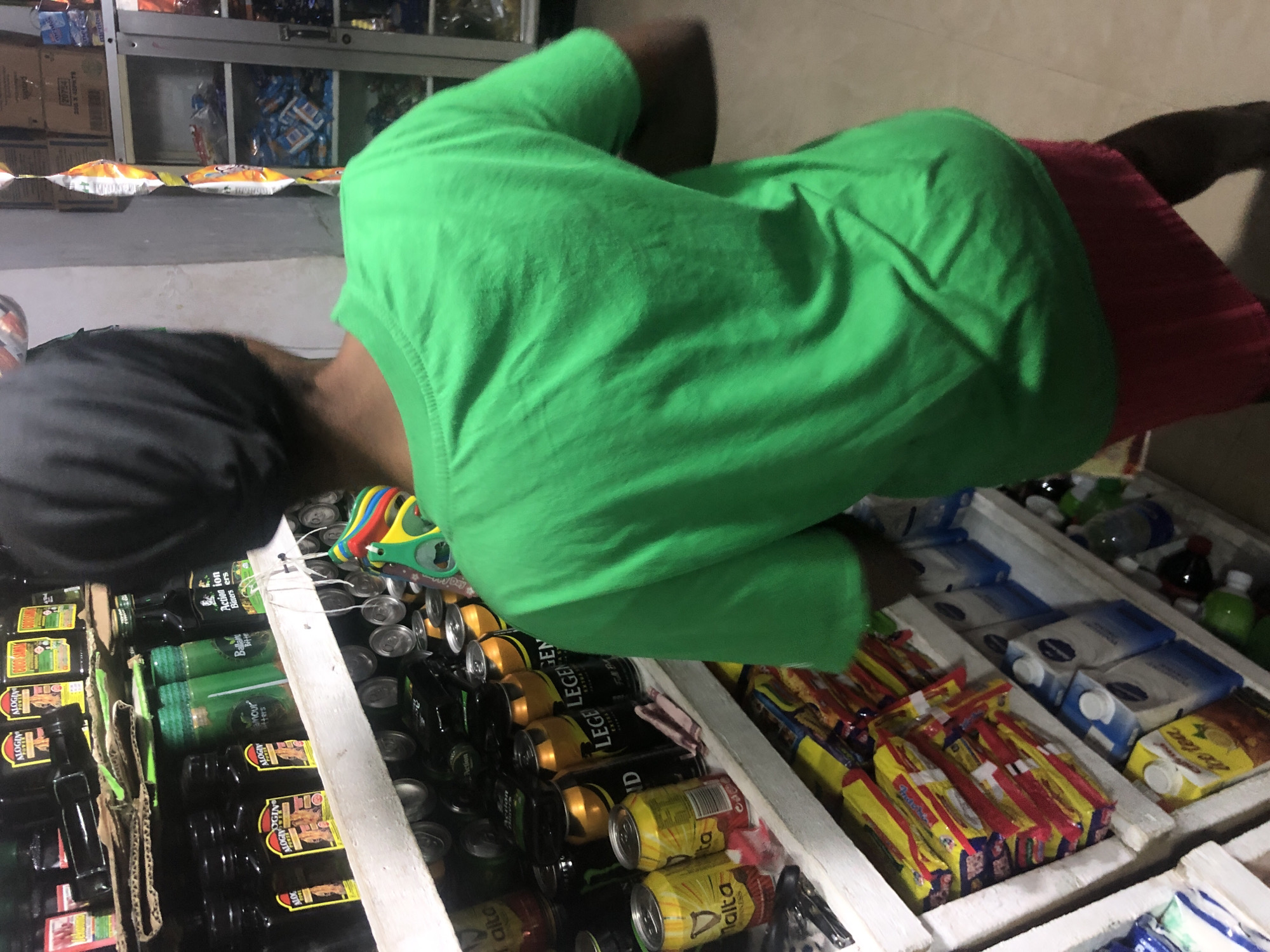 A person wearing a green t-shirt and a red skirt stands in front of shelves stocked with various products in a store in Ado, Nigeria. The shelves display an assortment of beverages, including cans of Malta Guinness, Legend Extra Stout, and Action Bitters. Packaged snacks and other bottled drinks are also visible. The person is partially obscuring some of the merchandise.