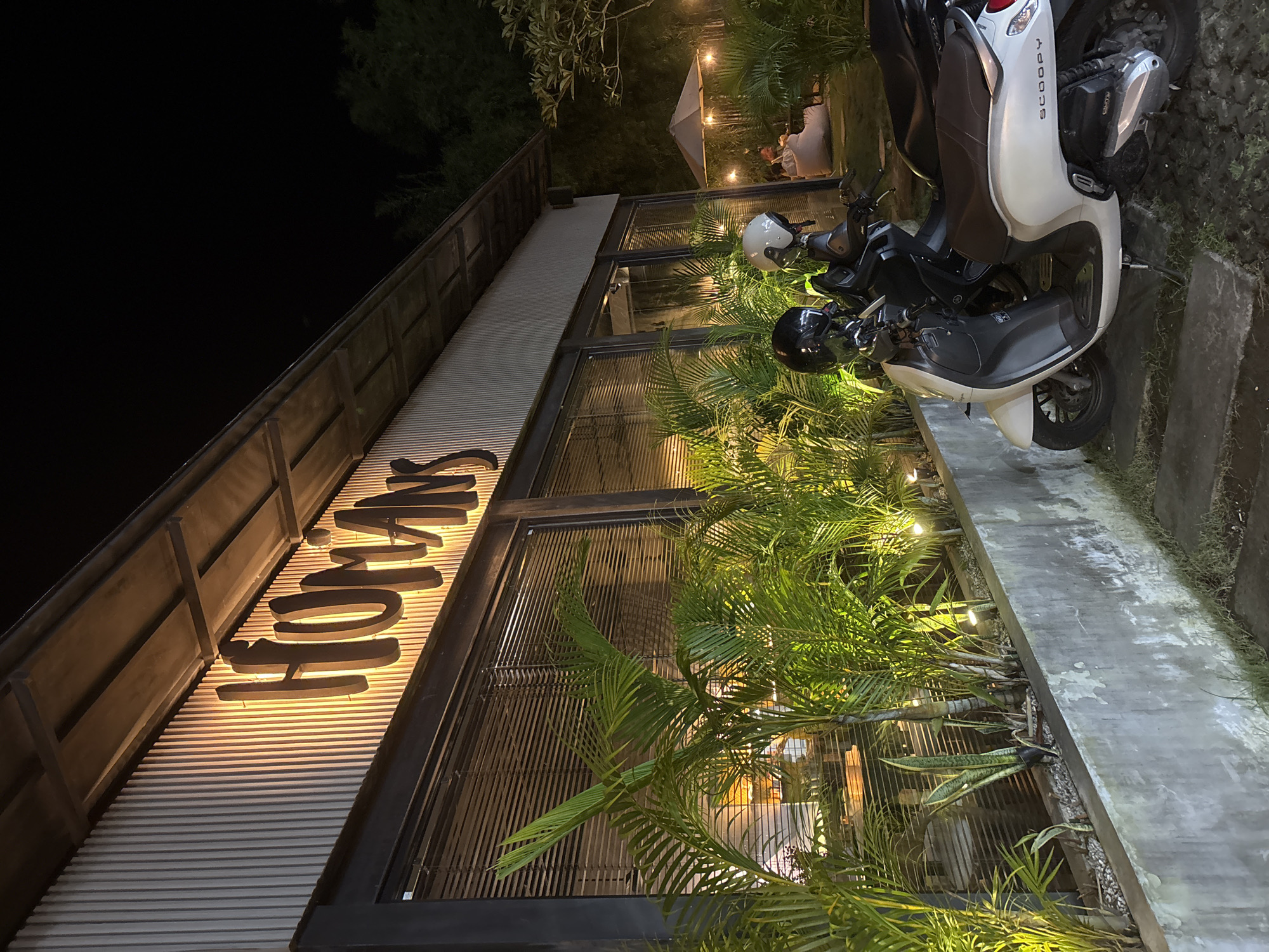 At night in Ungasan, Indonesia, a tilted eye-level shot captures a restaurant called "Homans." The facade features horizontal corrugated siding, with the restaurant's name in bold, illuminated letters. Large windows reflect the interior lighting and surrounding vegetation. A concrete walkway sits adjacent to the building. Two motorbikes are parked close to the entrance. A black helmet rests on the seat of the first motorbike, while a white helmet sits on the seat of the second. Palm trees and other tropical plants grow in front of the building. In the background, there is a seating area with a tent-like structure and people seated. The night sky is visible above the building.