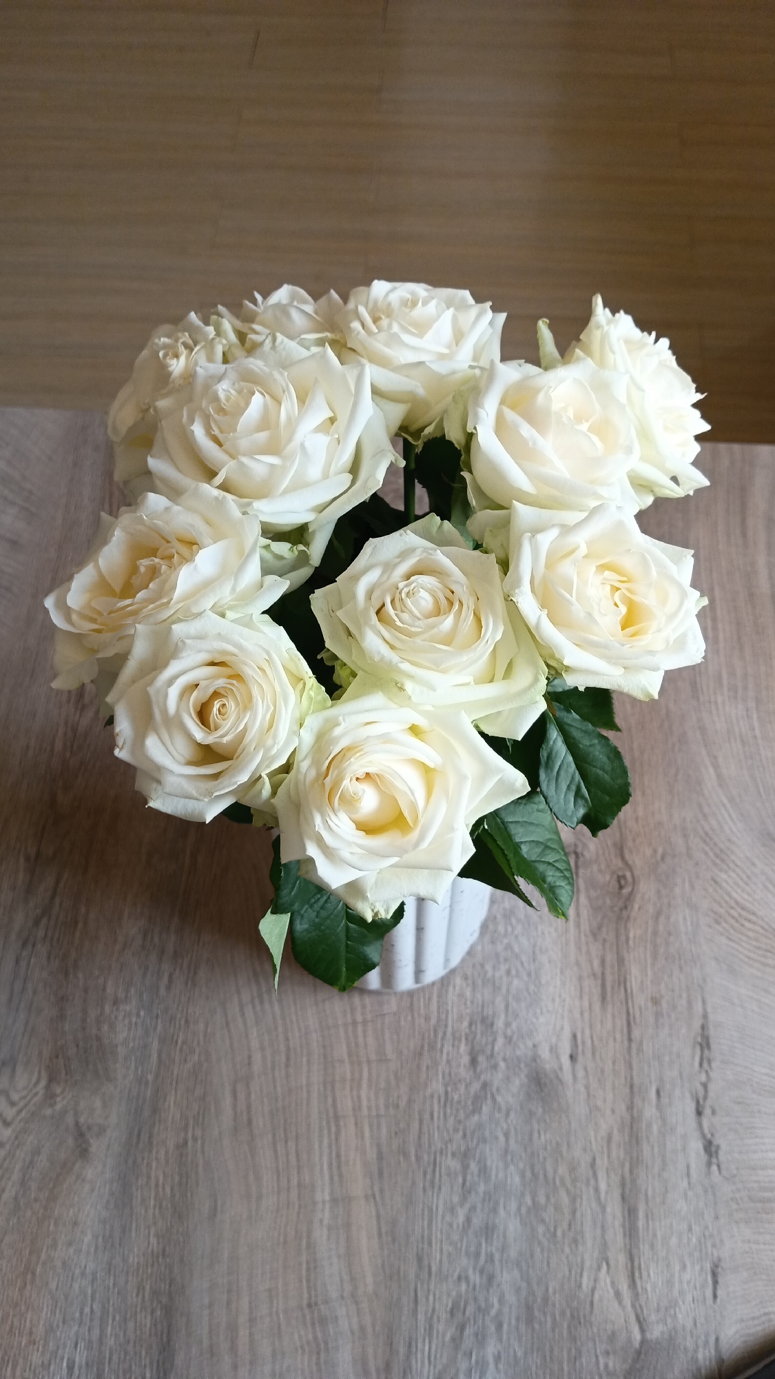 A bouquet of white roses is arranged in a white vase and sits on a wooden table. The roses are various stages of bloom, with some fully open and others still tightly budded. Green leaves are visible amongst the flowers. The background is a wooden surface with a horizontal grain. The scene is indoors.