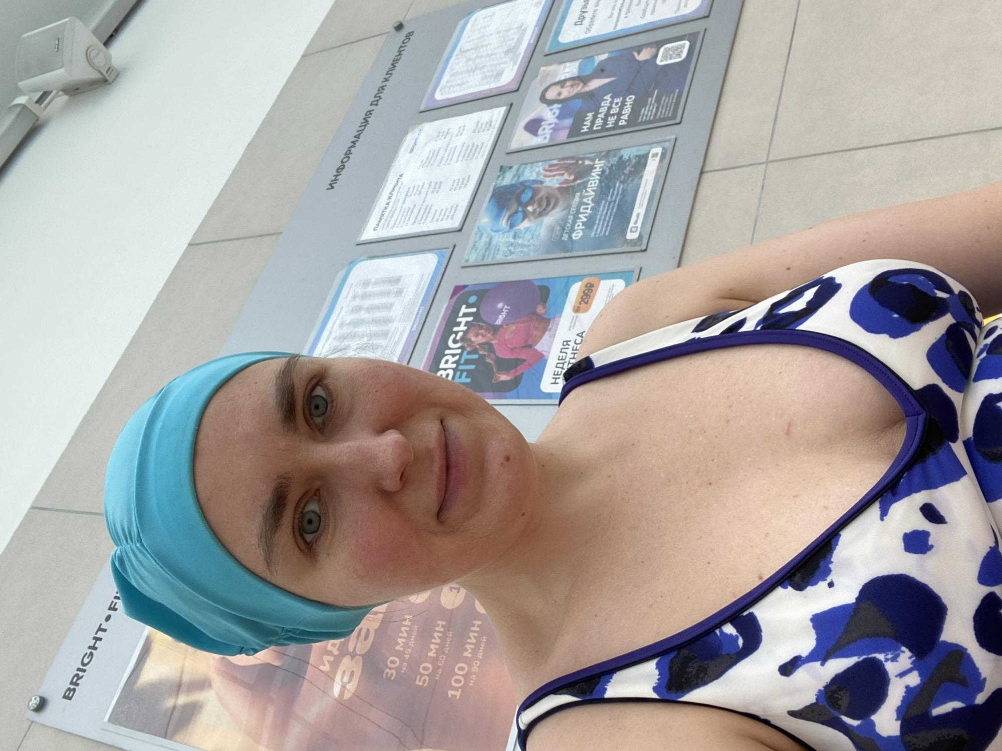 The image shows a woman wearing a blue swim cap and a blue and white patterned swimsuit. She is standing in front of a wall with several posters and information boards. The posters appear to be advertising a fitness or swimming facility, with text in Russian visible on some of them, including "BRIGHT FIT" and "ИНФОРМАЦИЯ ДЛЯ КЛИЕНТОВ". One poster features a woman in athletic wear, another shows a person snorkeling, and a third displays a woman in a suit. The woman in the foreground is looking at the camera and smiling slightly. The lighting suggests an indoor environment, possibly a public pool area. Without further context or visible landmarks, it is impossible to definitively determine the specific location in Yekaterinburg, Russia, though the posters suggest a commercial establishment.