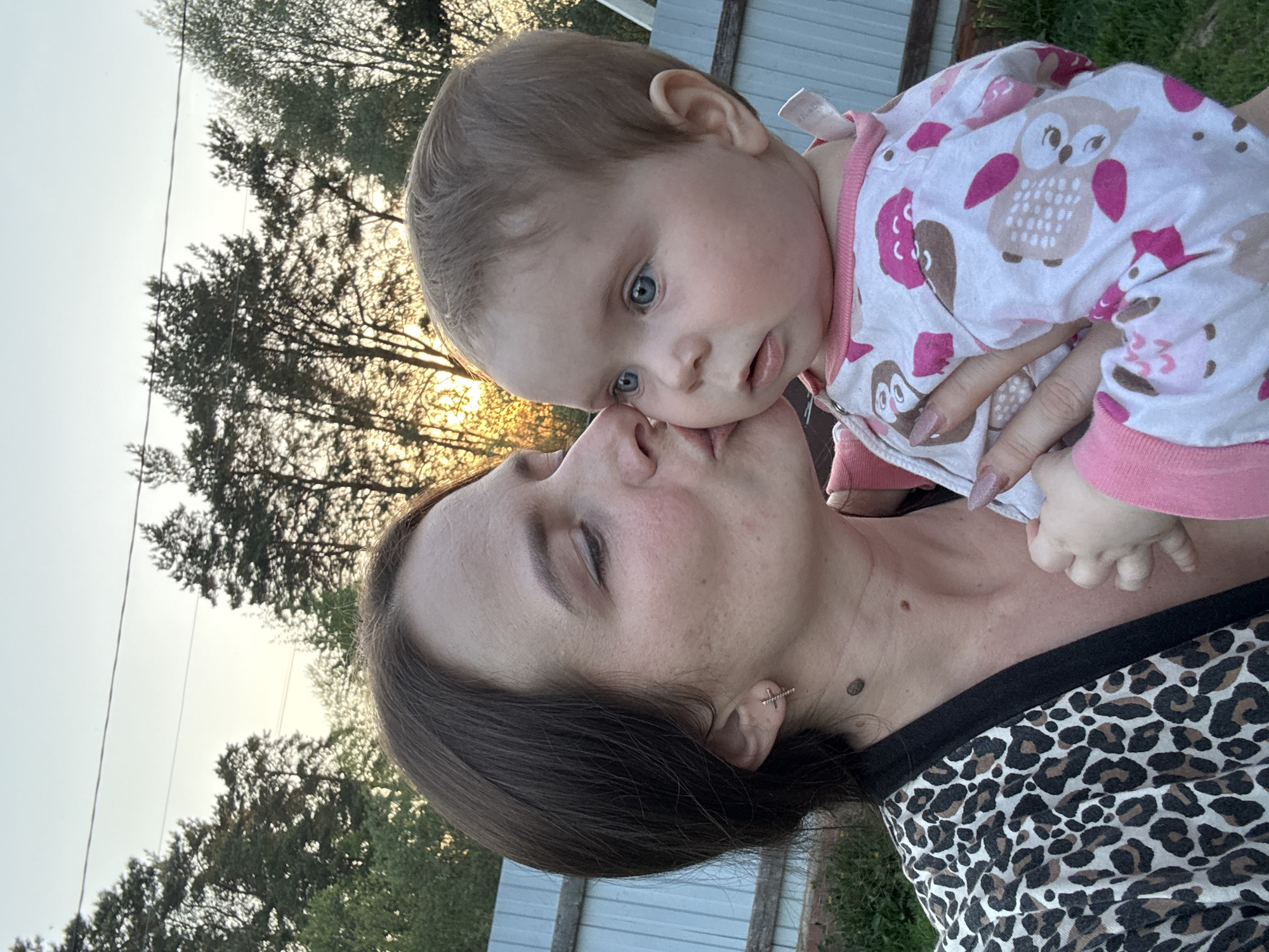 A woman with dark hair, wearing a leopard print top and a small earring, is holding a baby. The baby, dressed in owl-patterned pajamas with pink accents, is looking upwards with blue eyes and has their face close to the woman's face. The background shows trees and a partially visible structure, possibly a fence or building. The location is Rusalevka, Russia.