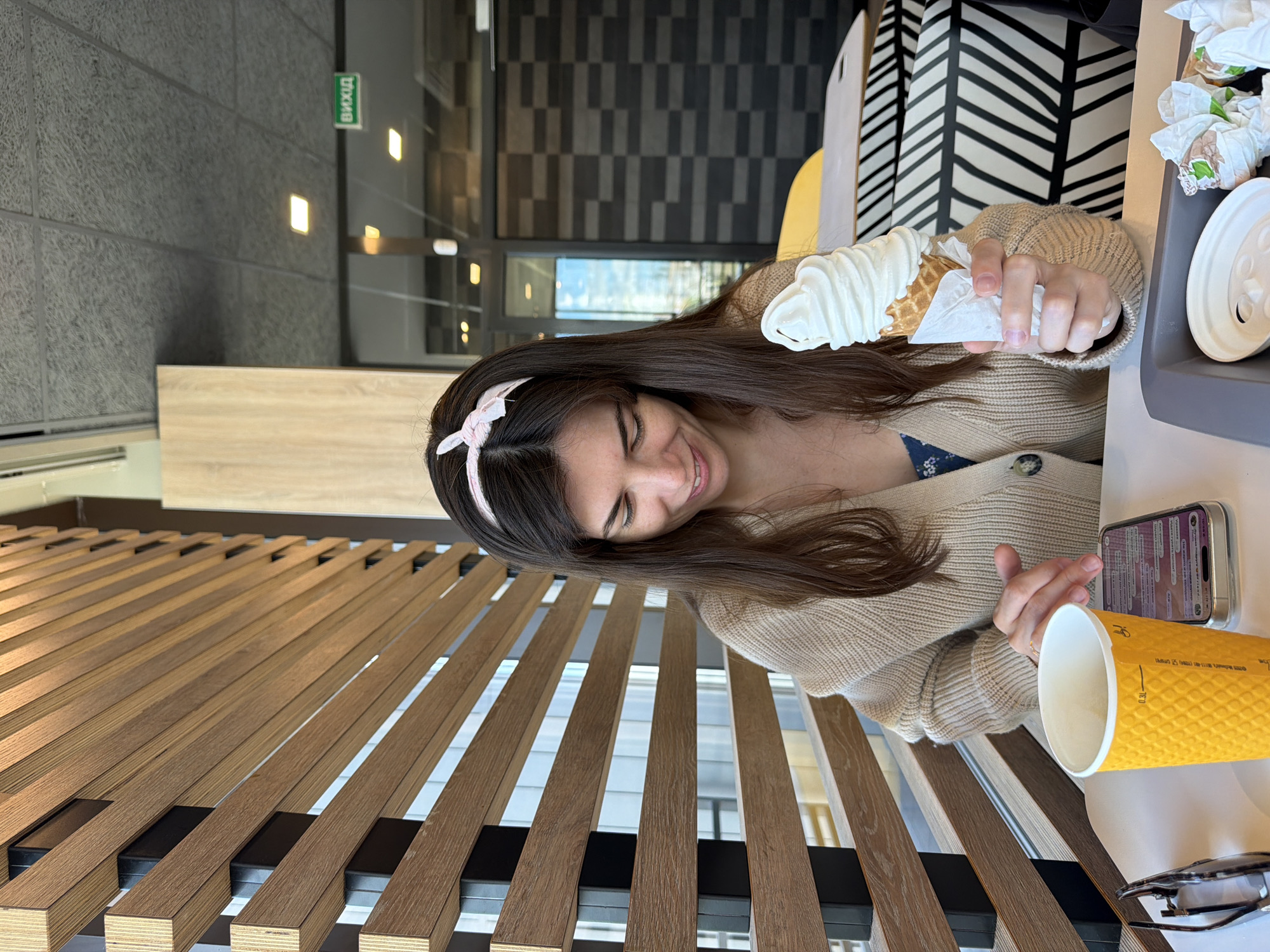 A woman is sitting at a table in a McDonald's restaurant in Kyiv, Ukraine. She is holding a soft-serve ice cream cone and looking down at her phone. In front of her is a yellow cup and a phone. The restaurant has wooden slat walls and a grey acoustic ceiling. There is an exit sign in Ukrainian.