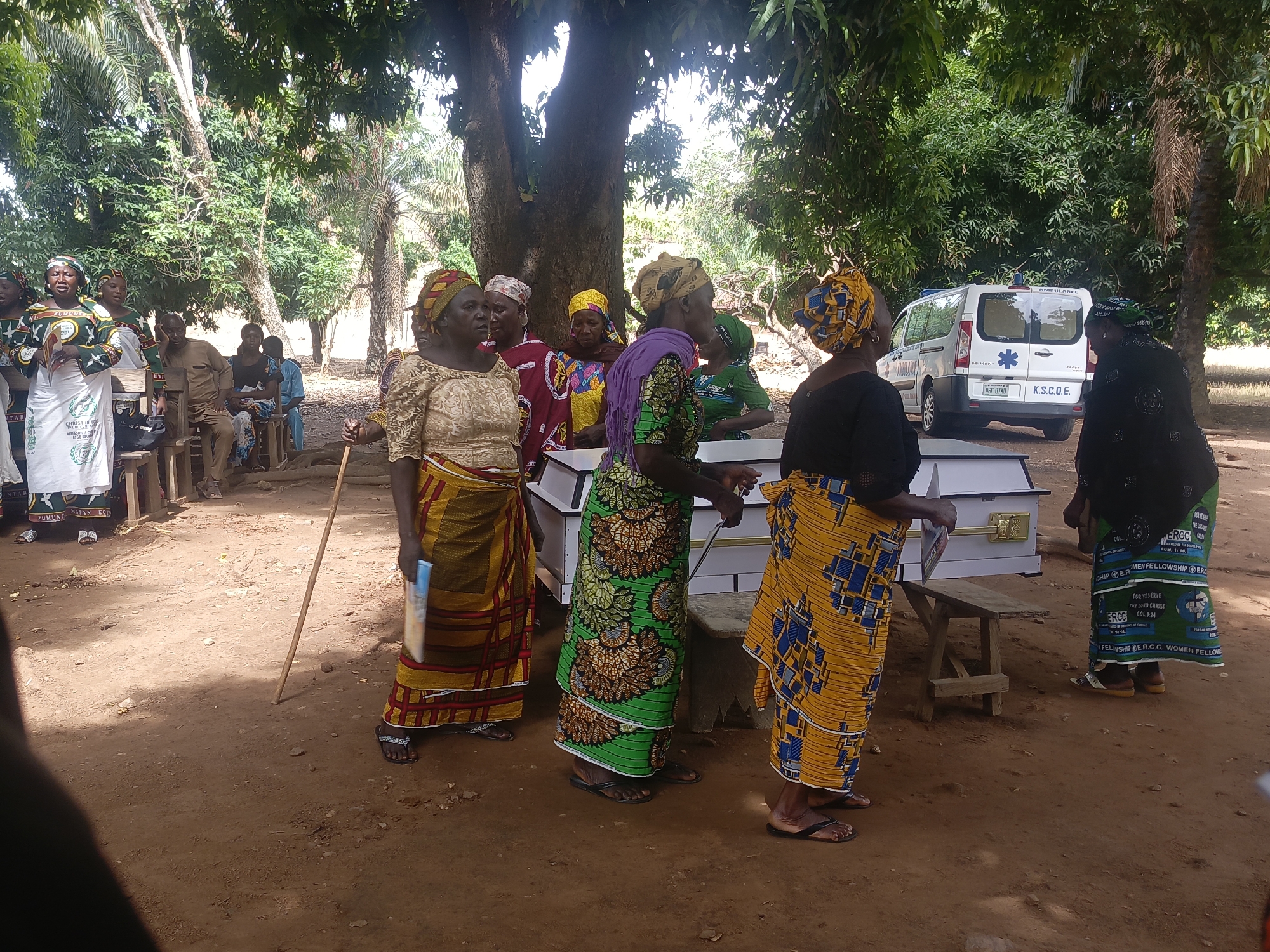 The image depicts a funeral scene, likely taking place outdoors in what appears to be a rural setting. A group of women dressed in traditional African clothing are gathered around a white coffin placed on a wooden stool. An ambulance is parked nearby, partially visible in the background. In the distance, more people are seated under the shade of trees. The ground is reddish-brown soil. Text on the garments of the women mentions "ZUMUNT MATAN EC," "MEN FELLOWSHIP EXC," "ERCC," and "WOMEN FELLOW." Based on the identification of "ERCC" (Evangelical Reformed Church of Christ) and the location of the image, it is likely the scene is taking place in or near Ninzo South, Nigeria.