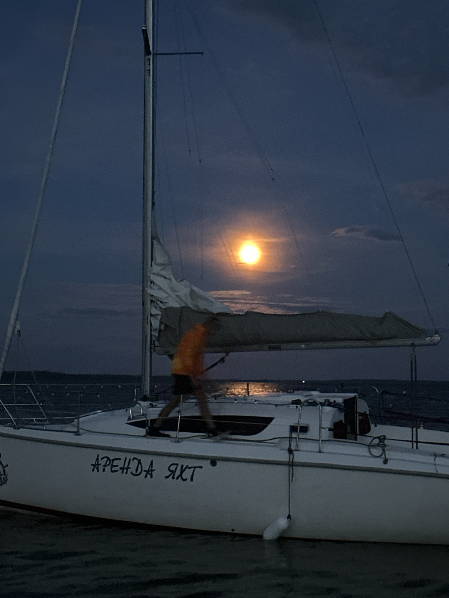 The image depicts a sailboat docked on the Naroch River in the Narochsky rural council of Belarus during twilight. The moon is visible in the dark blue sky, casting a soft glow on the scene. A person, blurred due to motion, is standing on the deck of the sailboat, wearing an orange shirt and dark shorts. The sailboat has the Cyrillic text "АРЕНДА ЯХТ" (Rent a Yacht) painted on its white hull. The water is calm, reflecting the moonlight. The surrounding environment appears to be a body of water with a distant dark shoreline.