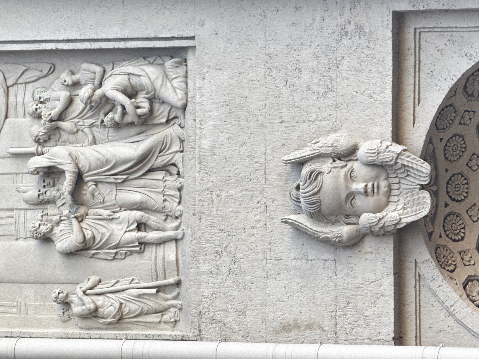 The image shows a close-up of architectural decorations on a building exterior, likely located in Saint Petersburg, Russia. The building's façade is made of a light-colored, textured material, possibly stone or concrete.
On the left side of the frame is a rectangular relief panel depicting a scene with multiple figures in classical robes and poses. The arrangement of bodies suggests themes of death, suffering, or a lamentation.
To the right of the relief panel is a protruding sculpture of an angel's head with wings. The angel's face is serene. Adjacent to the angel sculpture is a portion of a round, decorative element.
The scene is viewed from a slightly low angle, and a horizontal pipe or molding runs along the bottom edge of the frame.