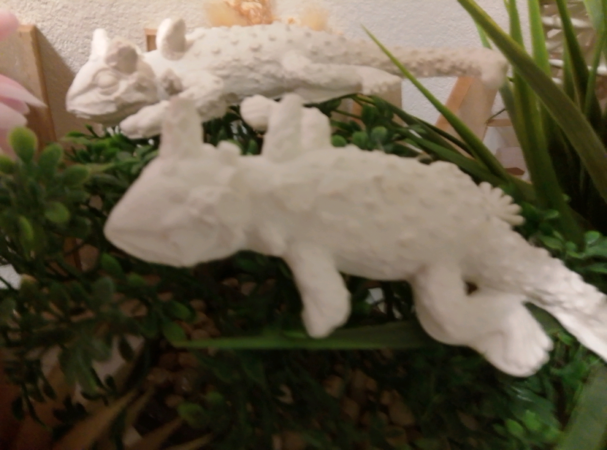 The image shows two white ceramic lizard figurines placed on a bed of artificial greenery. The figurine in the foreground is larger and positioned horizontally, facing left. It has visible legs, a tail, and a detailed textured body. The second figurine, positioned behind and above the first, is also white and appears to be a lizard in a similar posture, but it is partially obscured and out of focus. The figurines are surrounded by dense green artificial foliage and some small, pebble-like objects. A wooden element, possibly part of a shelf or decorative structure, is visible to the left and behind the figurines. A blurry pink object, likely a decorative flower, is also present to the far left. The background is a textured wall. The image was taken indoors, in a setting that appears to be a display or decorative arrangement. There is no indication of the city of Wedel, Germany.