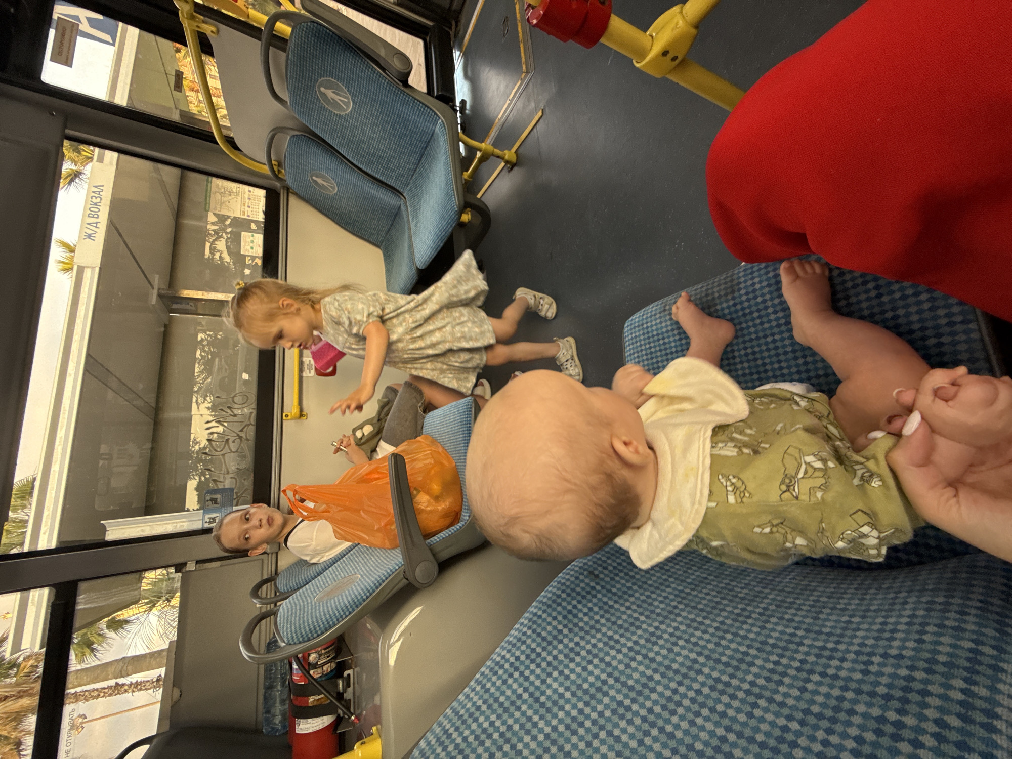 Inside a public bus, a baby wearing a light green outfit with a frog print sits on a blue patterned seat, facing away from the camera. The baby's head is tilted to the right. To the left, a pre-teen girl with a light complexion and a white shirt sits on a similar blue seat, looking to her left with an orange bag in her lap. In the aisle, a toddler in a light-colored dress is captured mid-motion, appearing to dance or move. The bus interior features blue seats with yellow framing and a gray floor. Outside the window, palm trees and a sign reading "Ж/Д ВОКЗАЛ" (Railway Station) suggest a location in Sochi, Russia. A red fabric is visible in the foreground.