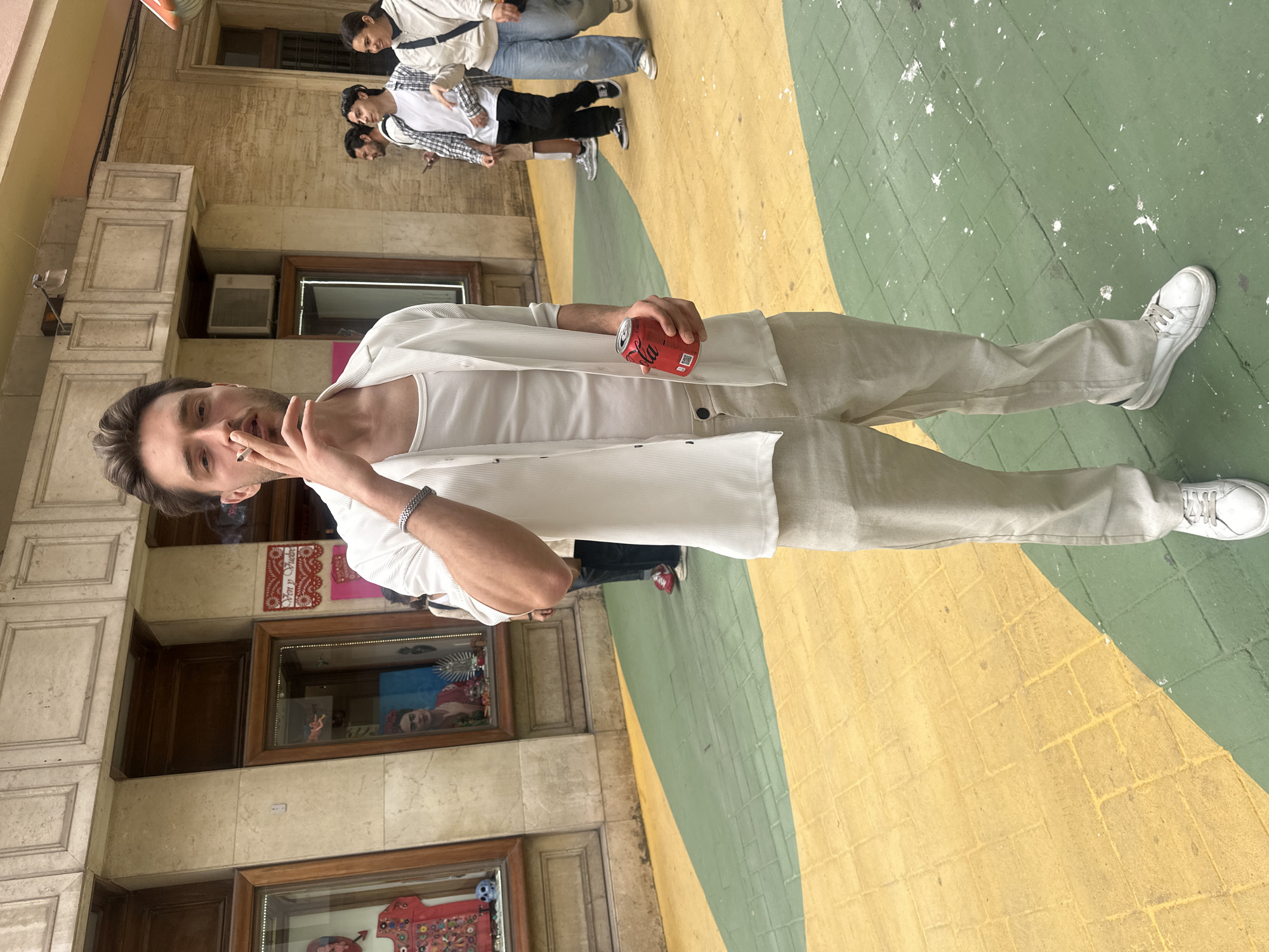 The photograph captures a young man standing outdoors in what appears to be a vibrant urban setting in Alicante/Alacant, Spain. The man, positioned in the foreground, looks directly at the camera while holding a lit cigarette to his mouth with his left hand and a red Coca-Cola can in his right hand. He is dressed in a white ribbed tank top, an unbuttoned white long-sleeved shirt with subtle stripes, light beige pleated trousers, and white sneakers. A silver chain bracelet is visible on his left wrist.
The ground beneath him is distinctly paved and painted in large, contrasting sections of yellow and green, with scattered white paint splatters. In the background, several other people are visible, some walking and others standing. One person wears a white jacket and blue jeans, while another has a plaid shirt over a white T-shirt and black pants.
Behind the figures are buildings with light-colored, possibly stone or stucco, facades featuring ornate window frames and wooden elements. Through a shop window or display to the man's right, colorful decorations are visible, including red and white paper cutouts with partial Spanish text that seems to say "VEN Y PINTA" (Come and paint) and an image resembling Frida Kahlo. The scene appears to be taking place during the daytime under soft lighting, suggesting either an overcast sky or a shaded area. The overall atmosphere is casual, with the main subject posing relaxed for the shot.