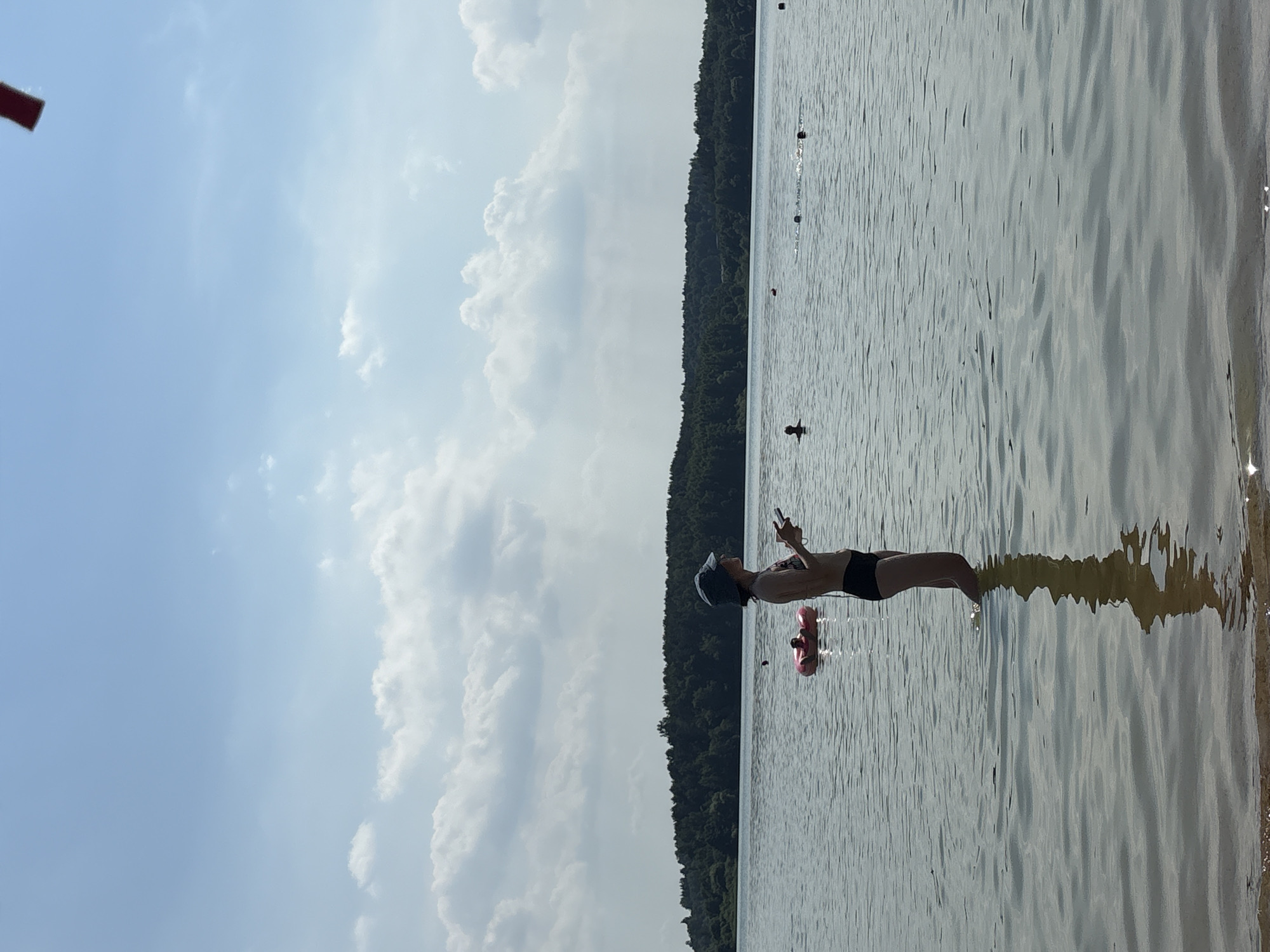 A wide shot captures a sunny day at a lake near Ozyaritska-Slabadski rural council, Belarus. In the foreground, a person wearing a bucket hat and a bikini stands knee-deep in the calm water, holding a phone. Their reflection is visible on the water's surface. Further out in the lake, a pink inflatable ring with a person inside floats, and other individuals are swimming. The background features a dense, dark green treeline bordering the lake, with a bright blue sky filled with scattered white clouds above. A small red object is visible at the very top edge of the frame.