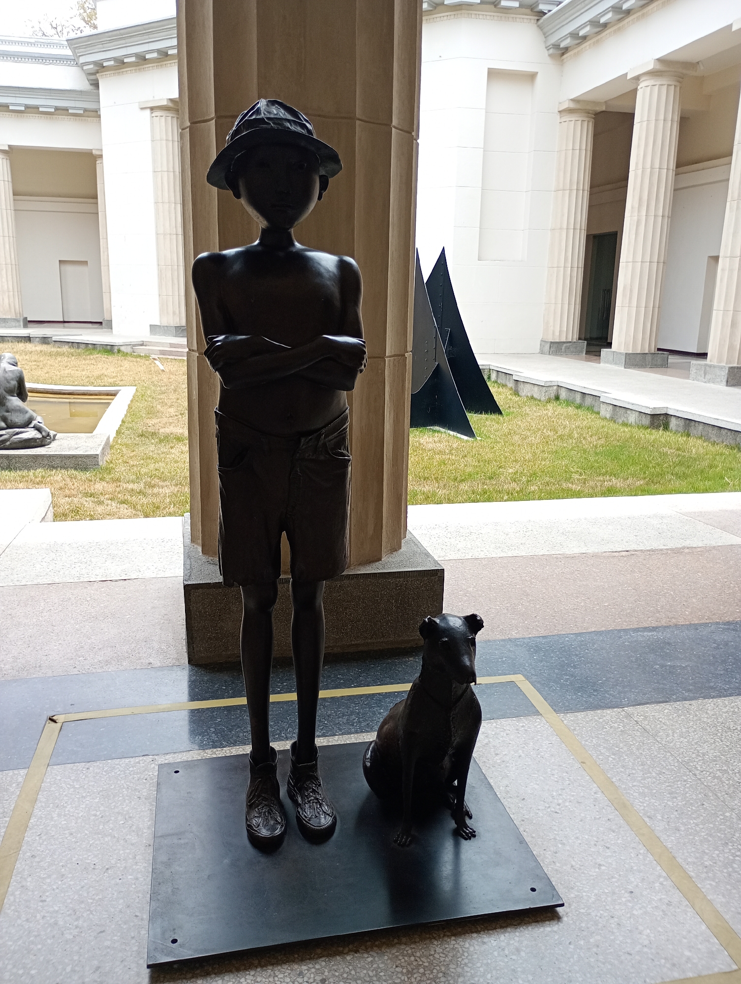 This image captures a bronze sculpture of a young boy with his dog, situated in what appears to be an outdoor gallery or museum courtyard in Caracas, Venezuela. The main subject, a dark bronze statue of a boy, stands centrally, facing forward with his arms crossed over his chest. He wears a wide-brimmed hat, shorts, and lace-up shoes. His face is smooth and stylized, without detailed features. To his right, a bronze statue of a dog sits attentively on the same dark, rectangular base, looking in the same direction as the boy.
The statues are placed on a patterned tiled floor, featuring grey and beige sections with prominent yellow demarcation lines. The immediate surroundings suggest a covered walkway or portico, characterized by large, fluted, light-colored columns that flank the central sculpture. Beyond this covered area, an open courtyard features a well-maintained lawn.
In the background, elegant, classical-style buildings with multiple columns and cornices are visible, suggesting a significant public or cultural institution. On the left side of the grass, another smaller, partially obscured statue (possibly a kneeling figure) is seen next to a shallow water feature or pond. On the right, a large, black, angular abstract sculpture stands in the grass. The scene is well-lit by natural daylight, indicating daytime, with some shadows suggesting the time of day or the covered nature of the walkway. No visible text is present in the image. The overall impression is one of art displayed within a grand architectural setting.
