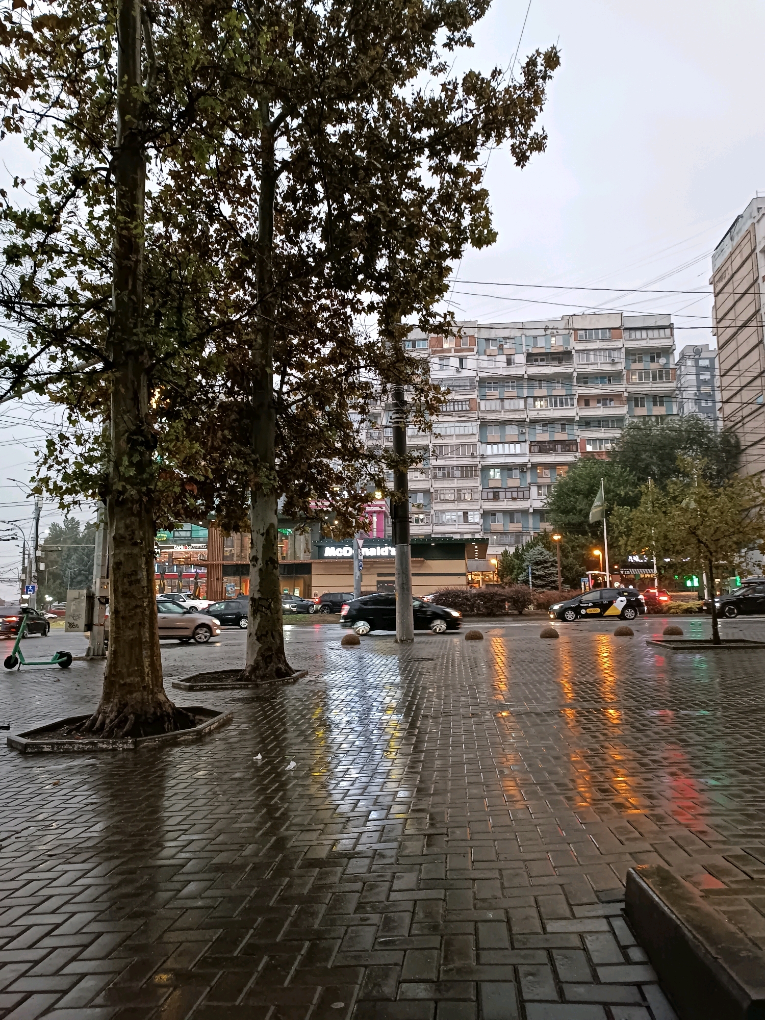 This image depicts a street scene in Chișinău, Moldova, on a wet day. In the foreground, two large trees with autumn foliage stand on a wet, patterned brick sidewalk. The pavement reflects the lights of passing vehicles and surrounding buildings, indicating a damp environment.
Behind the trees, a McDonald's restaurant is visible, with its distinctive yellow arches and signage. Across the street, a multi-story residential building with numerous balconies dominates the background. The architecture suggests a Soviet-era construction style, common in many Eastern European cities.
Several cars are present on the street, including a light-colored sedan, a black SUV, and a yellow and black taxi with markings "101" and "Taxi 14700". A green electric scooter is parked on the sidewalk to the left of the trees. The sky is overcast, contributing to the muted light of the scene. Electrical wires are strung across the sky, typical of urban infrastructure. The overall impression is that of a transitional season, likely autumn, with the wet conditions adding a reflective quality to the urban landscape.