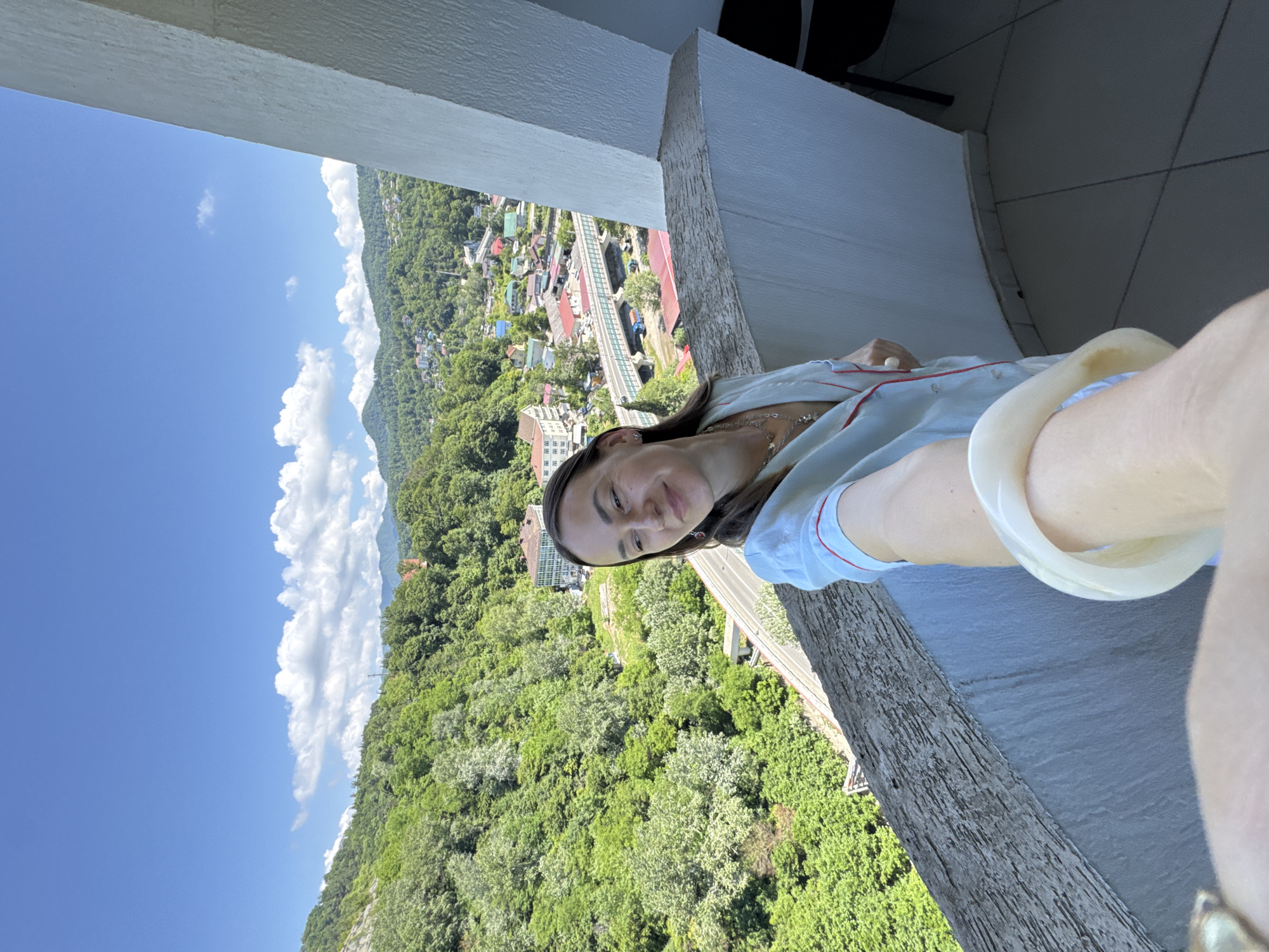 A woman stands on a balcony in Sochi, Russia, taking a selfie. The balcony has a textured railing and a white wall. The background features a lush green landscape with a town nestled in a valley. A bridge and road are visible, winding through the town. The sky is blue with scattered white clouds. The woman is wearing a light blue pajama set with red trim and several necklaces. She has a bracelet on her wrist. Her body is angled to the side as she looks into the camera. The foreground shows a portion of her arm and hand, holding the phone.