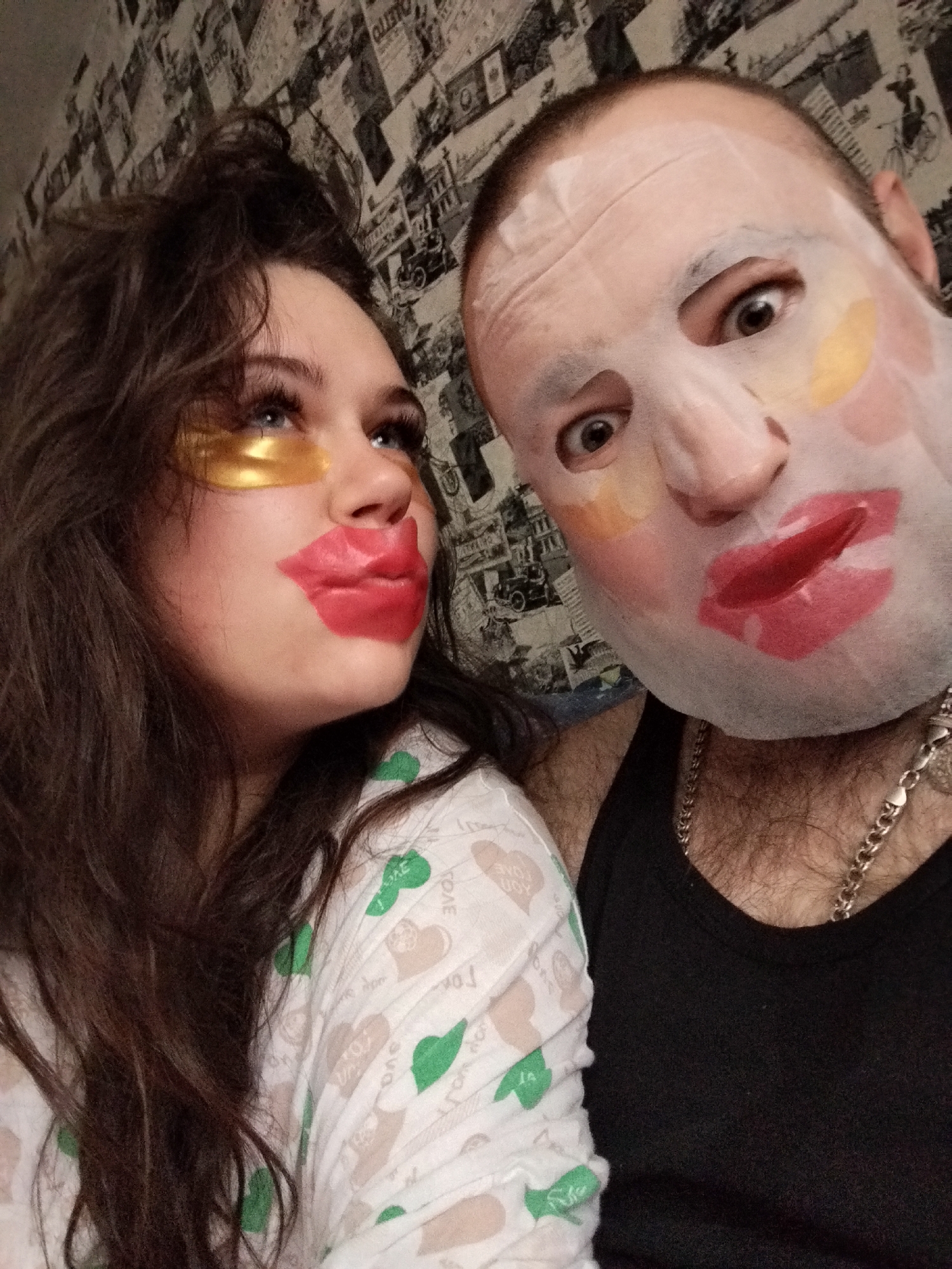 A selfie depicts a man and a woman, both wearing facial masks. The woman on the left wears golden under-eye patches and a red lip mask, has long wavy brown hair, and is wearing a light-colored top with green and brown motifs. The man on the right wears a full facial mask with a red lip overlay and other colored sections, a black tank top, and a silver chain necklace. The background shows a wallpapered wall with monochrome urban scenes, and appears to be an interior setting, likely a home. The location is Chaikovsky, Russia.