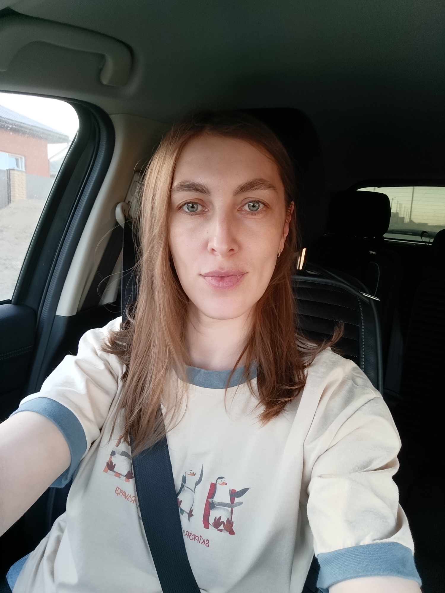 A woman is in the driver's seat of a car. She is wearing a t-shirt with an illustration of penguins and a seatbelt. The car is in Chaikovsky, Russia, as evidenced by the building visible through the window. The interior of the car is black.