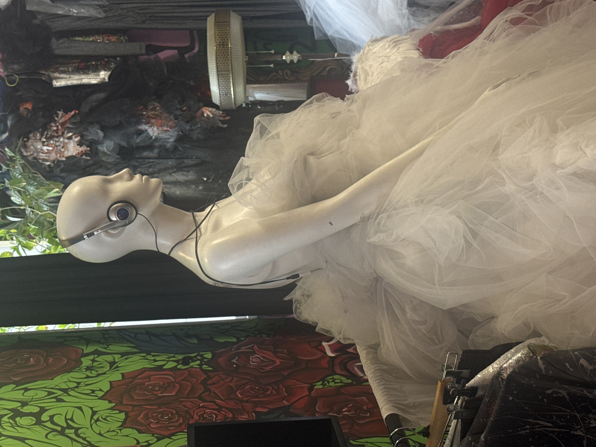 A mannequin wearing headphones and a white tulle dress is displayed in what appears to be a retail store. The mannequin is positioned to the right of a wall adorned with a vibrant green backdrop featuring large red roses. Behind the mannequin, dark fabric drapes obscure the background, with hints of other clothing items and a lamp visible. The overall scene suggests a boutique setting, possibly in Komsomolsk-on-Amur, Russia, given the context of the request, though the image itself provides no direct evidence of its location.