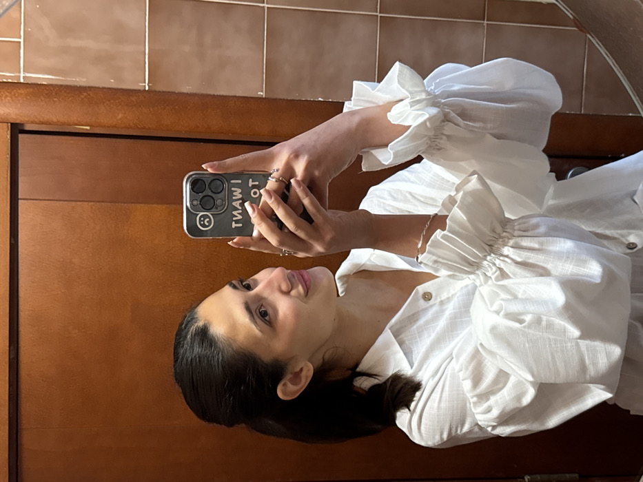 A female individual is depicted in a mirror selfie, captured indoors in Budva, Montenegro. The female, appearing in her 20s or 30s, has dark hair pulled back into a ponytail and is smiling. She wears a white, collared shirt featuring prominent ruffled, elasticized cuffs on long, voluminous sleeves. Her fingernails are painted with light polish. She wears multiple rings on both hands and a twisted silver-toned bracelet on her left wrist. She is holding a silver-colored smartphone, equipped with a triple-lens camera system, in her right hand to take the photograph. The smartphone is encased in a clear cover displaying an inverted sad face emoji and the inverted text "I WANT TO". The background behind her left shoulder features a light brown wooden door, while light brown square wall tiles are visible behind her right shoulder.