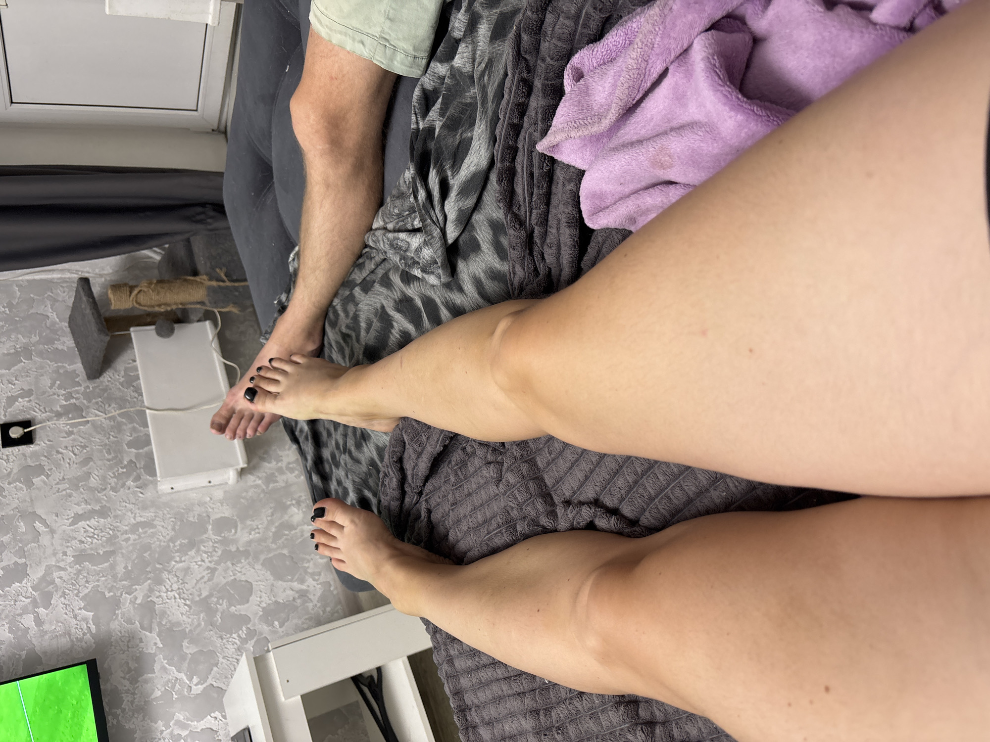 The image shows a close-up view of two pairs of legs resting on a dark gray blanket. On the left, a person's legs are bare from the knees down, with visible skin texture and faint shadows. Their toenails are painted black. To the right, a person's legs are also bare from the knees down. They are wearing light green shorts. Their right leg is bent at the knee. A purple folded towel is visible near the bottom right of the image. In the background, a wall with textured wallpaper and a white shelf are visible. A television screen displaying a green field is partially visible in the upper left corner. A cat tree is visible in the upper right corner. The image appears to be taken indoors. The location in the city of Tolyatti, Russia is not specified.