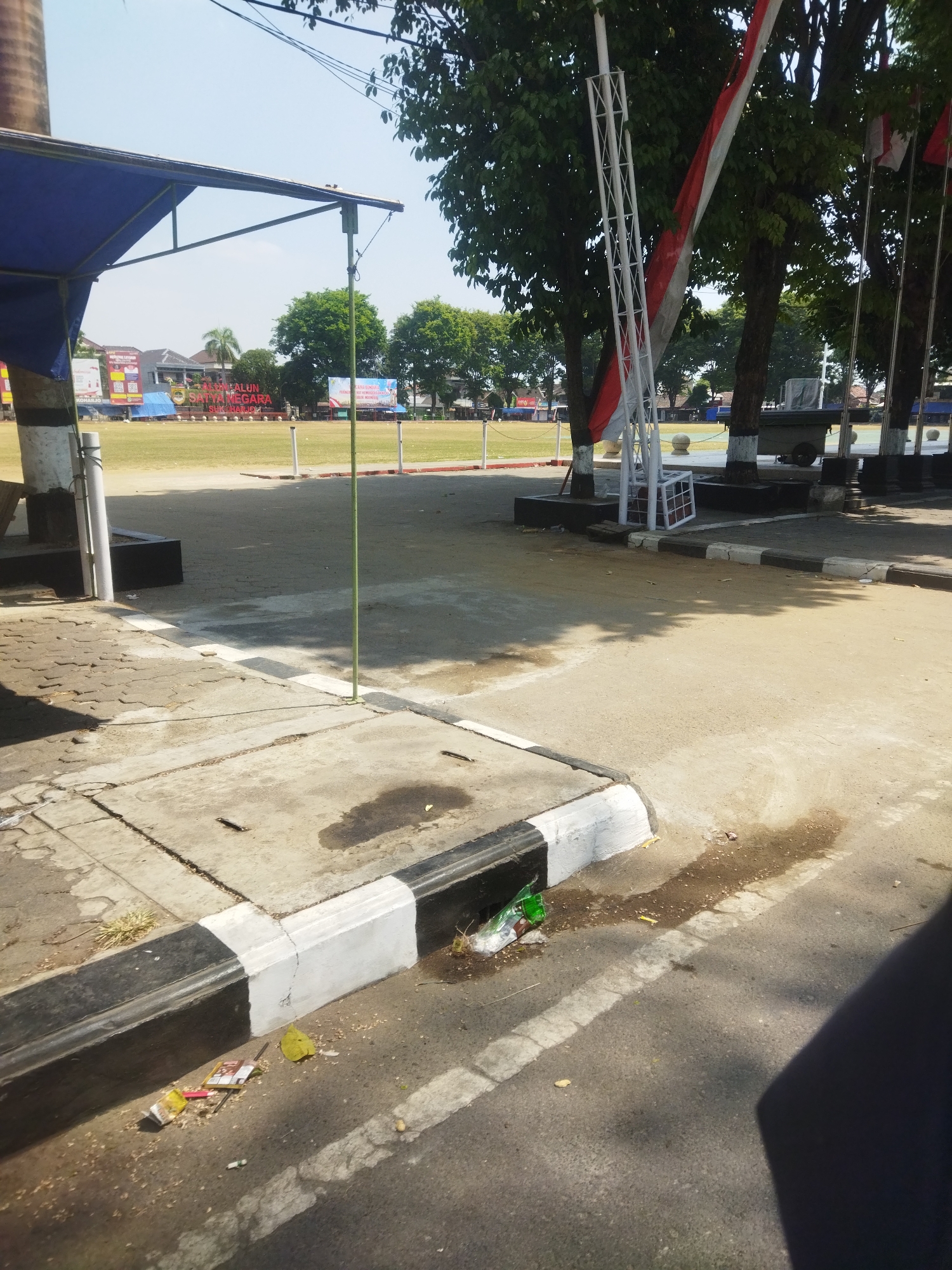 The image shows an outdoor scene on a sunny day, likely in Sukoharjo, Indonesia, with a large open field in the background. In the foreground, a paved area with a curb is visible, which is partially covered in debris including a green plastic bottle, cigarette packs, and scattered leaves. The curb is painted with alternating black and white sections. Adjacent to the paved area is a sidewalk made of concrete blocks.
In the mid-ground, a large field with some tents and banners is visible. A prominent banner with red and white text, possibly a slogan or announcement, is placed on the field. Behind the field, there are buildings and trees.
To the right of the field, a tall metal structure, possibly part of a stage or a flagpole, stands near some trees. Indonesian flags are visible on flagpoles behind the metal structure and the trees. The trees are lush green and appear to be mature. The overall environment suggests a public gathering or event space. The ground shows signs of recent rainfall or spillage, with dark wet patches on the asphalt.