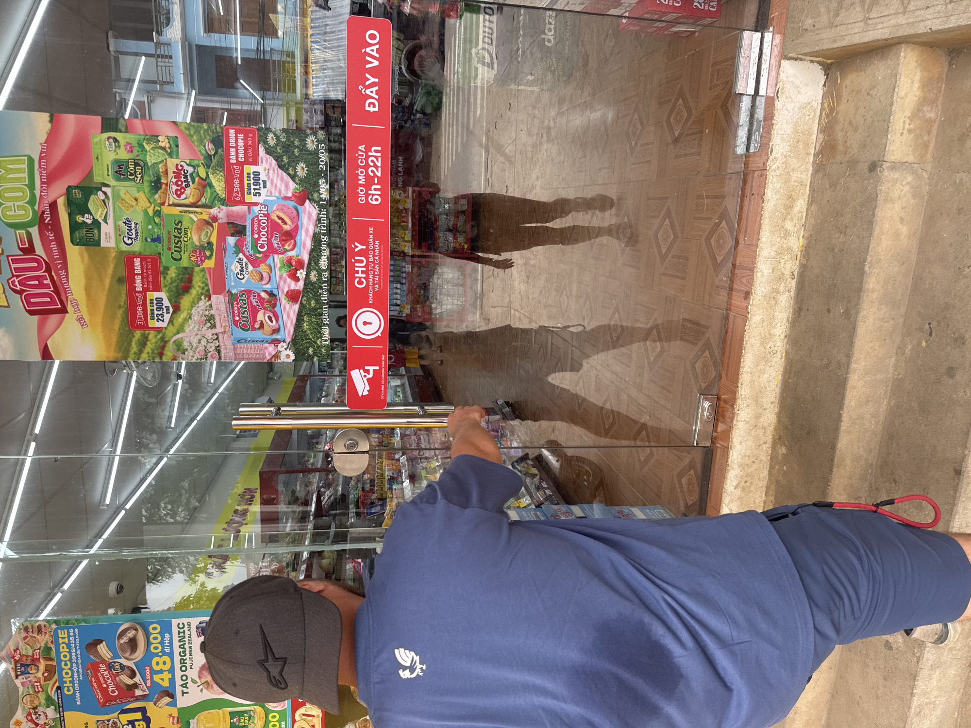 A person wearing a blue t-shirt and shorts, and a grey baseball cap, stands in front of a glass door, reaching for the handle. The person's back is to the camera. The door is the entrance to a store in Bắc Yên, Vietnam. The store appears to be a convenience store or supermarket, with shelves stocked with various products visible through the glass. A red sign with white text is affixed to the door, indicating store hours and a notice about security cameras. The floor inside the store is tiled. Outside the store, there are concrete steps leading up to the entrance. The overall lighting suggests it is daytime.