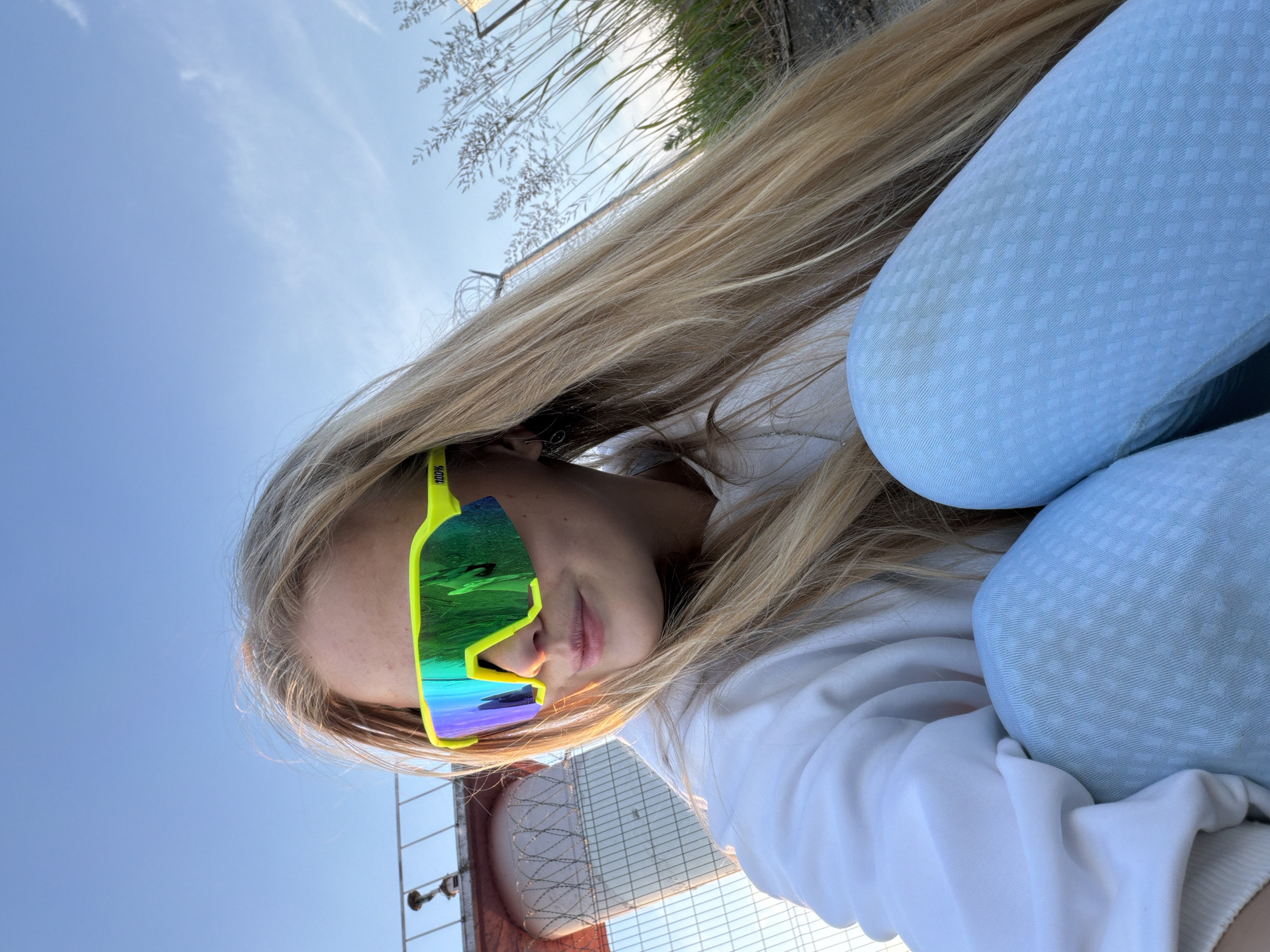 A young person with long blonde hair is sitting outdoors, looking to their left. They are wearing a white hoodie and light blue patterned leggings. Their eyes are obscured by large, neon yellow wraparound sunglasses with colorful mirrored lenses. In the background, there is a clear blue sky with a few wispy clouds. To the left, a chain-link fence and a portion of a red structure are visible. To the right, tall dry grass and plants grow. The ground appears to be rough, possibly asphalt or dirt. The lighting suggests it is late afternoon or early evening. The location is identified as Chaikovsky, Russia.
