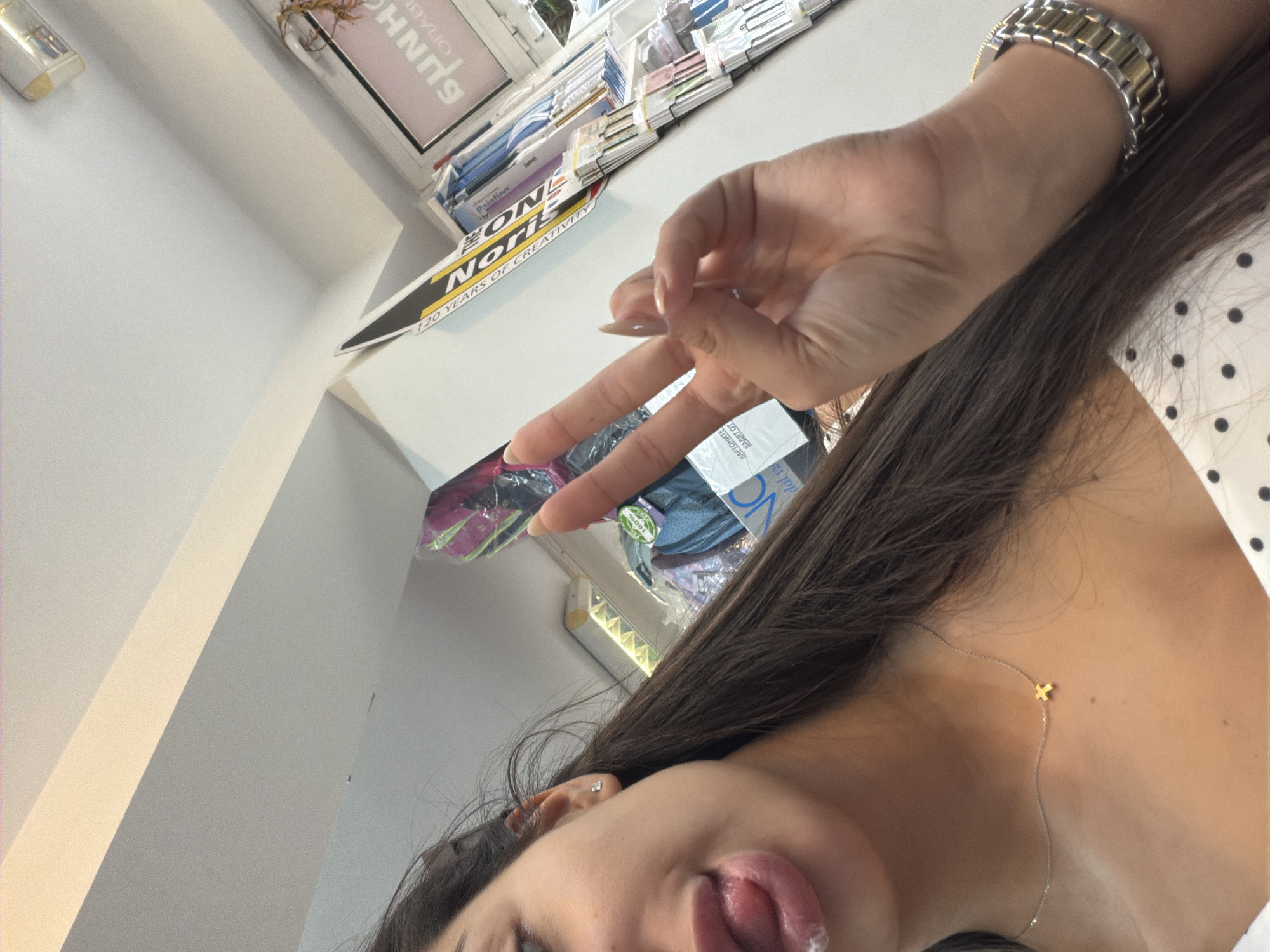 Here is a detailed description of the image:
**People:** The image features a woman. Her hair is dark, and she is wearing a watch and a necklace. She is sticking out her tongue and making a peace sign with her fingers.
**Objects:** Visible objects include a watch, necklace, and various papers or brochures with some branding ("Lone Note," "120 Years of Creativity," "SUNH").
**Environment:** The setting seems to be indoors, possibly a store or office.
**Location in Plovdiv, Bulgaria:** Due to the lack of specific landmarks or signs indicating the city of Plovdiv, it is not possible to determine the exact location. There is no mention of "Plovdiv" written in the available text. The phrase "dal 12" may indicate it is an Italian-oriented establishment, however, such establishments can exist in any city including Plovdiv, Bulgaria.
**Action:** The main action is the woman making a peace sign while sticking her tongue out.