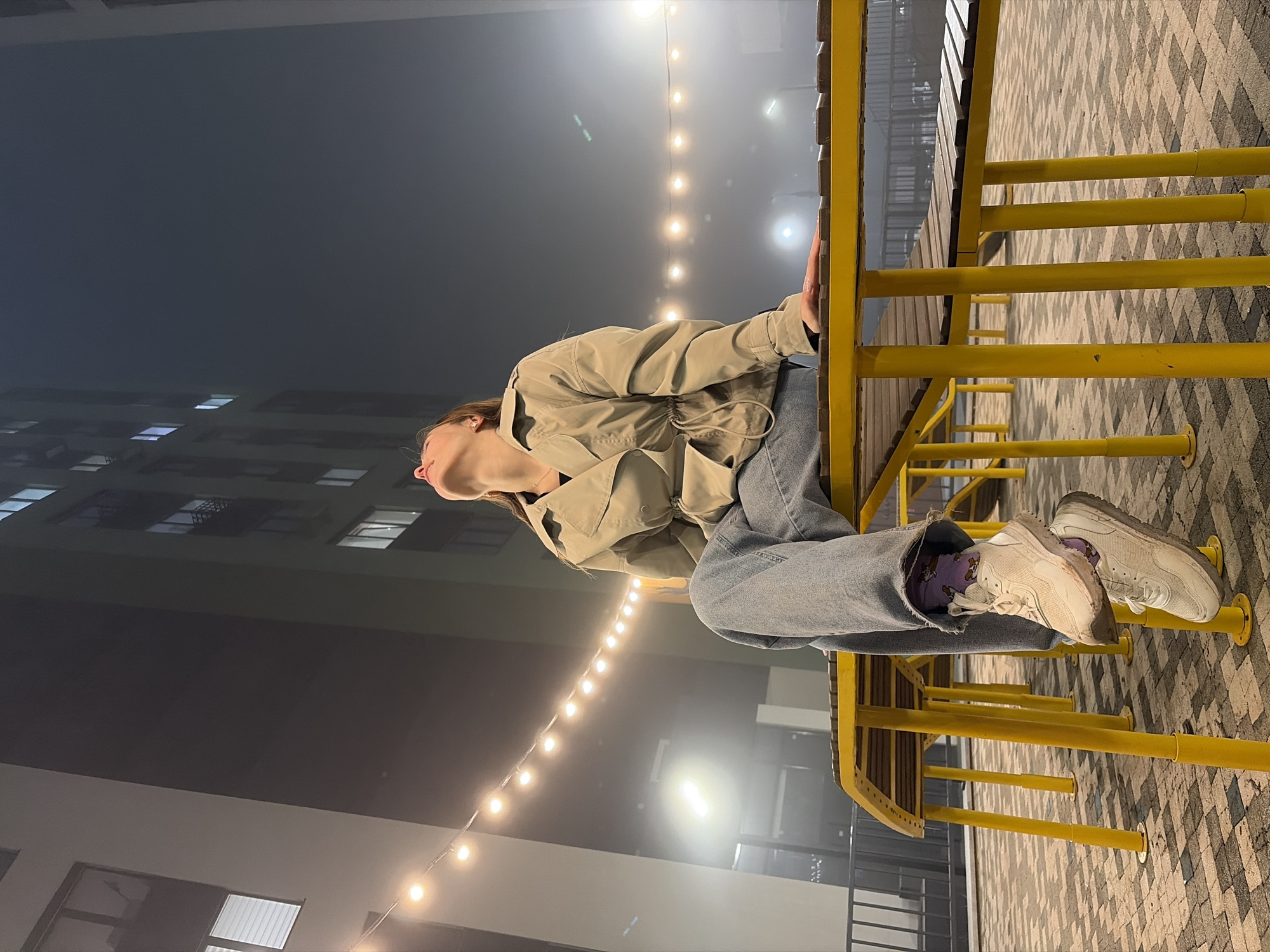 A person sits on a yellow structure in an urban setting at night. The individual wears a beige jacket and blue jeans with white sneakers, revealing purple socks. In the background, a tall building with illuminated windows is visible, as well as string lights hanging overhead. The location is Izhevsk, Russia.