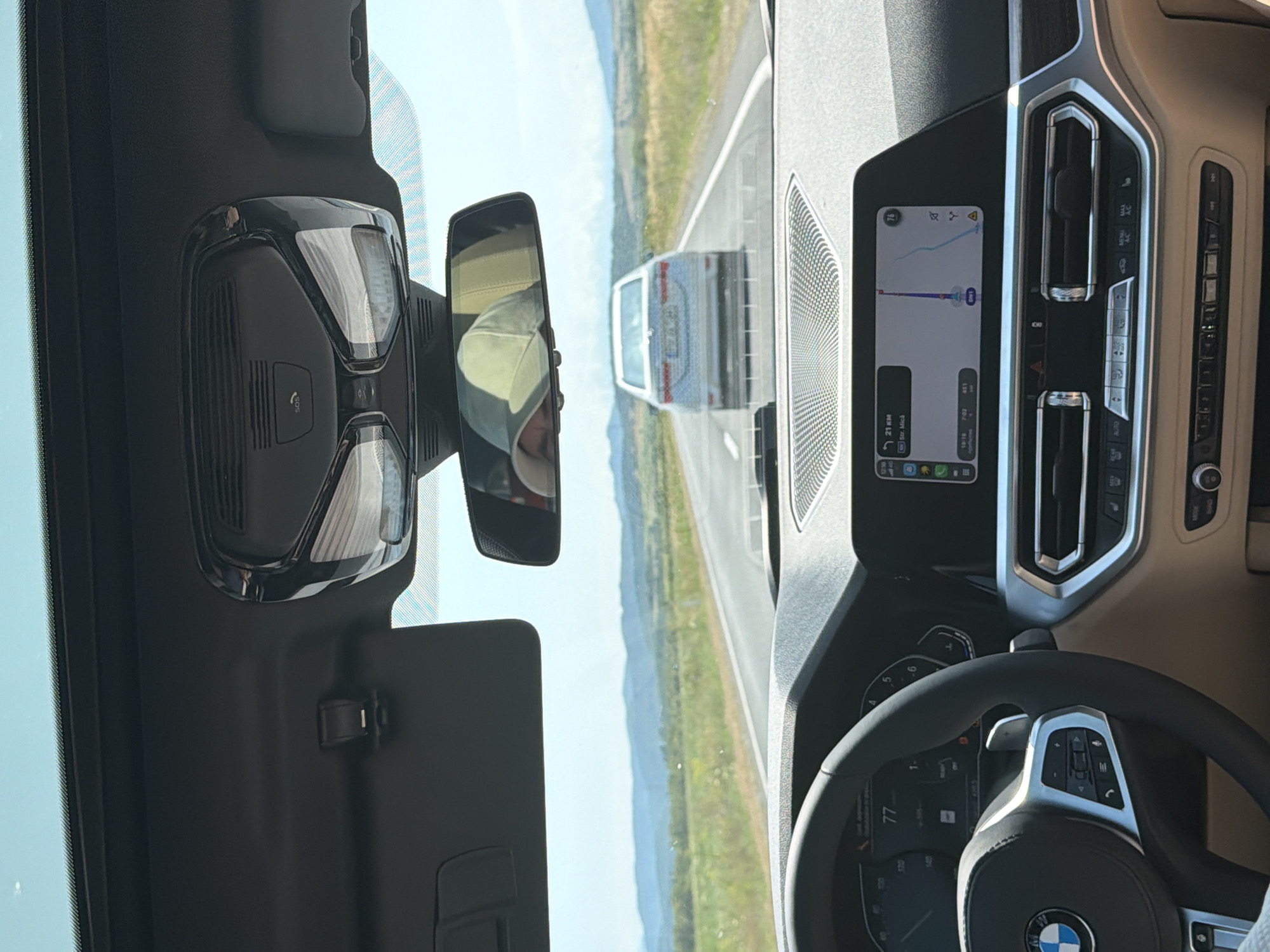 The image shows the interior of a car driving on a road near Perșani, Romania. The driver, wearing a light green cap, is visible in the rearview mirror. The car is following another silver sedan on a two-lane road bordered by grassy fields and distant hills. The dashboard displays navigation information with a route highlighted on a map. The sky is clear and blue.