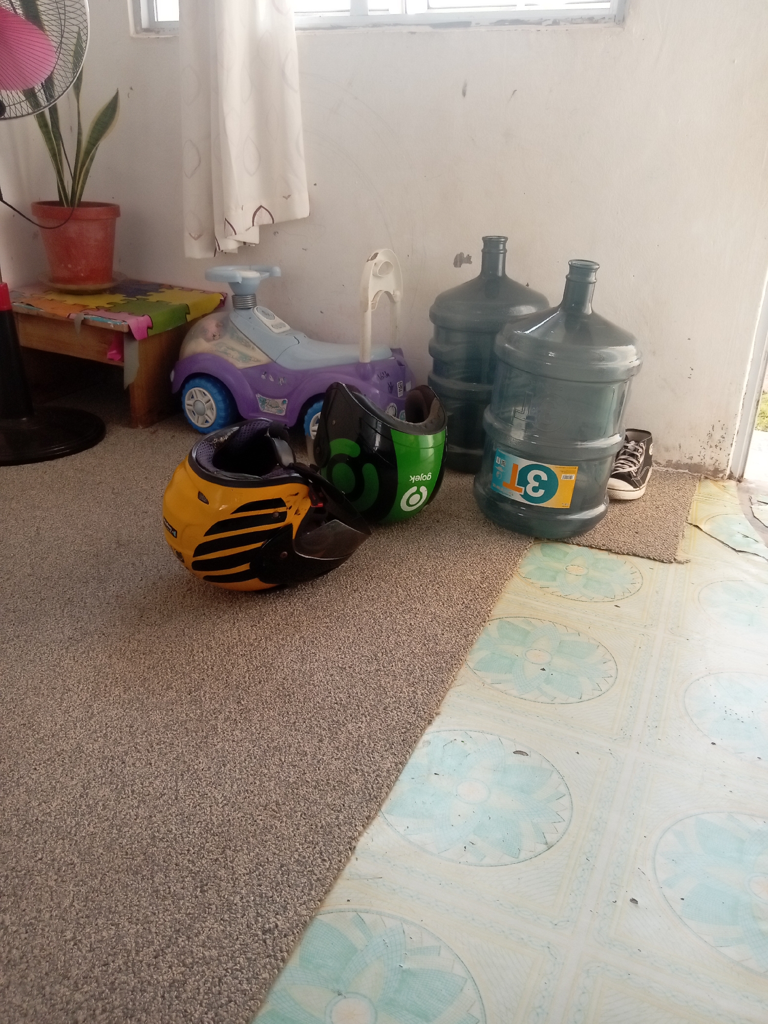 The image depicts an indoor scene with a neutral-toned carpet in the foreground and a tiled floor in the background. On the carpet, two helmets are visible: one yellow with black stripes and another green with black accents and the word "Gojek" printed on it. To the left of the helmets, a purple and blue ride-on toy car is positioned. Behind the toy car, a wooden table with a colorful puzzle mat is present. A potted snake plant sits on the table, and a pink-bladed fan is to its left. In the background, two large blue water jugs are stacked. A black and white sneaker is placed near the jugs. The overall setting appears to be a residential area in Batam, Indonesia, likely a living room or porch.