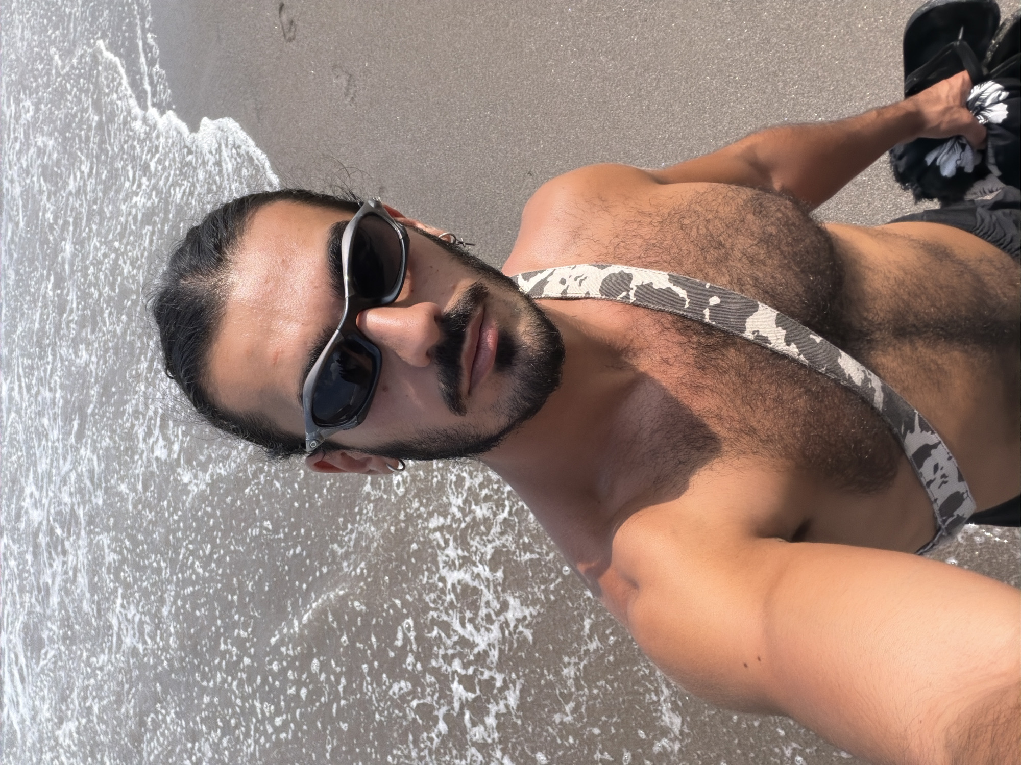 A man stands on a beach in North Kuta, Indonesia, with the ocean to his left and sand to his right. He is wearing sunglasses and a strap with a camouflage pattern across his chest. He is holding a bag in his right hand. The waves are breaking on the shore, and the sand appears wet where the water meets it. The man is looking at the camera, and his body is angled slightly to the right. The lighting suggests it is daytime, possibly midday, given the brightness and lack of long shadows.