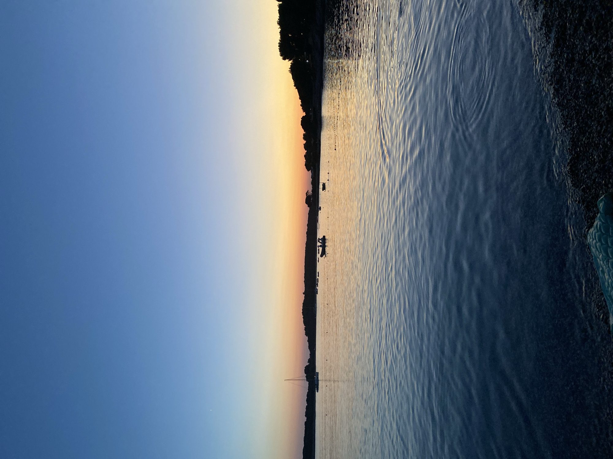 This is a vertical, eye-level, wide shot of the sea at dusk in Banjole, Croatia. The sky is a gradient of deep blue at the top fading to orange and yellow at the horizon. The horizon is marked by a silhouetted tree line. Several boats are visible on the calm water, including a sailboat with its mast extended and a small motorboat with people inside. The water is a deep blue, reflecting the colors of the sky. The foreground shows a pebbly shore with some green material visible at the bottom left. The overall atmosphere is serene and peaceful.