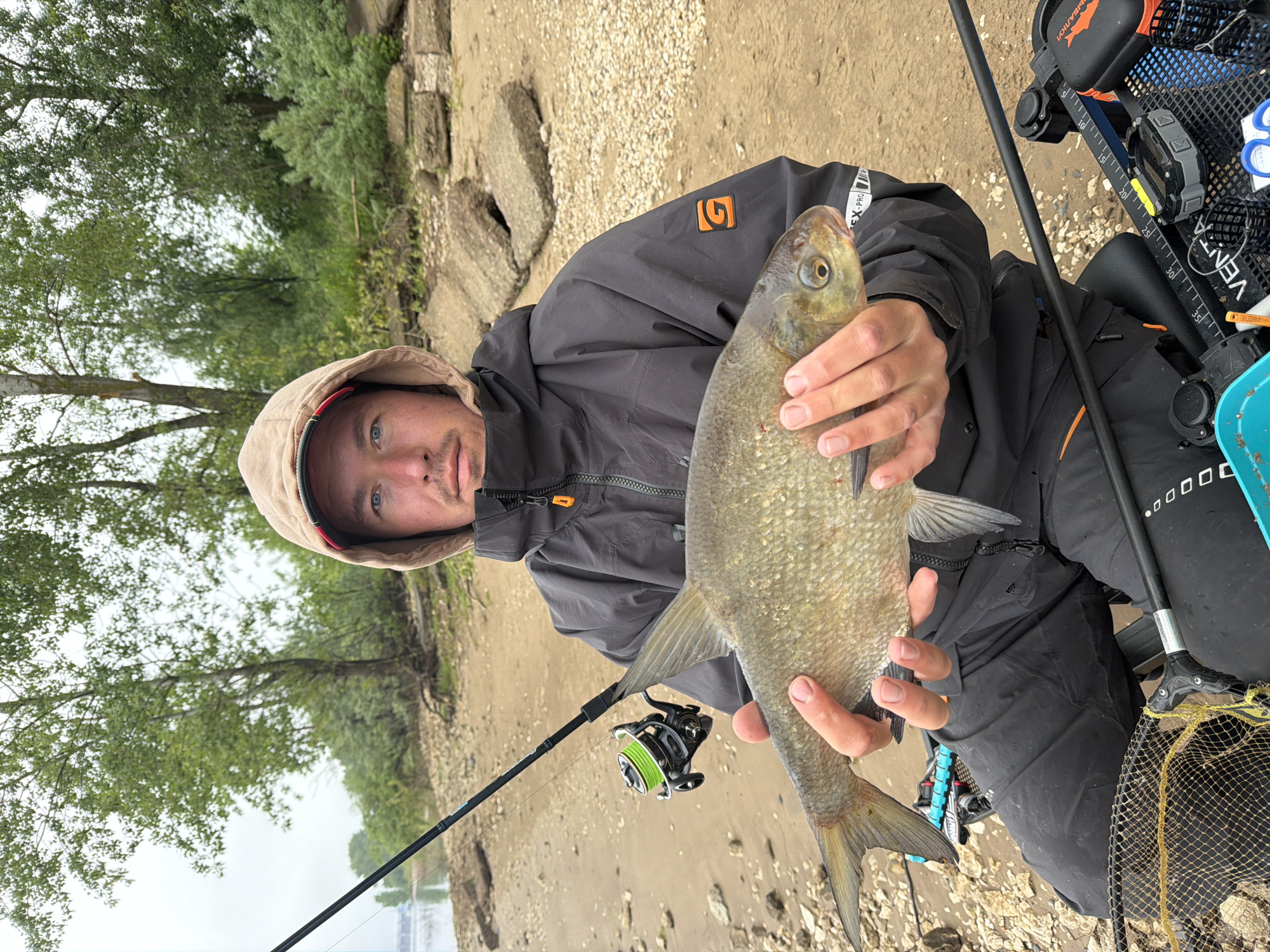 An outdoor portrait captured in Noviy, Russia, features a male angler holding a freshly caught fish. The man, with blue eyes and light facial stubble, is wearing a dark, hooded jacket with an orange 'G' logo on the sleeve and "EX-PRO" text partially visible. His light-colored hood is pulled up. He presents a medium-sized, silvery-green fish, possibly a bream, towards the camera, holding it horizontally with both hands.
The setting is a riverbank or lake shore, characterized by light brown sand and gravel, interspersed with larger rocks. A body of water is visible in the distance, and the opposite bank is lined with lush green trees and foliage under an overcast sky, suggesting a cloudy day.
Numerous pieces of fishing equipment surround the angler. A fishing rod with a black reel spooled with bright green line is propped nearby. To his right, a black fishing seat or box holds various tools: a black and yellow measuring tape with numbers like "30" and "35" clearly visible, orange-handled scissors, and a fishing net with a yellow mesh. Additional gear, including a tackle box with "SHAR..." (likely part of a brand name) and "VENTA" text, is also present. The man looks directly at the viewer with a neutral expression, indicating a successful fishing activity.