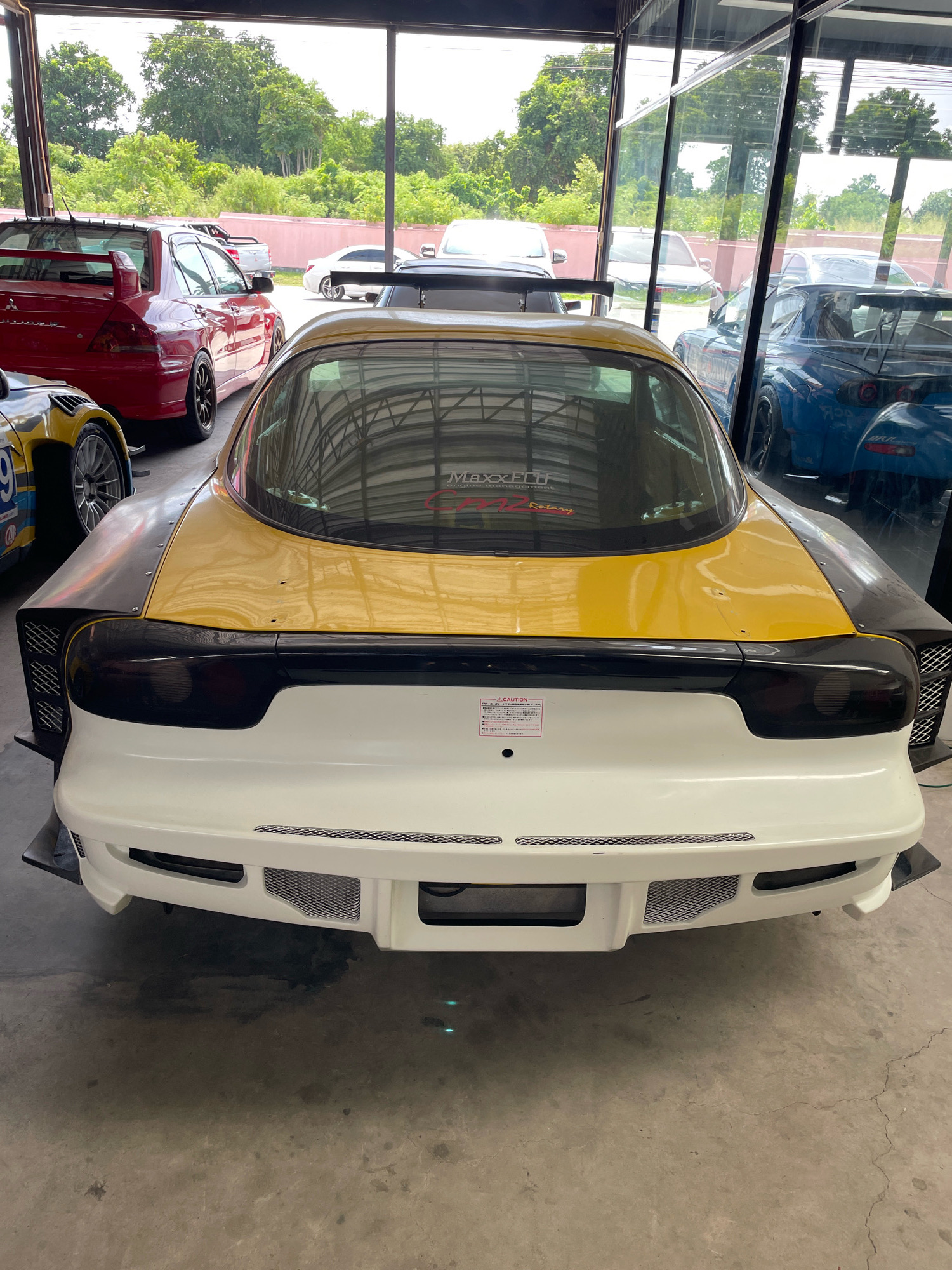 The image shows the rear view of a yellow and white modified Mazda RX-7 sports car parked indoors. The car has a prominent rear spoiler and wide body kit. In the background, through large glass windows, trees and other vehicles can be seen. One of the other cars visible is a red Mitsubishi Lancer Evolution. The setting appears to be a car showroom or garage in the city of Nong Prue, Thailand. The floor is concrete and the lighting suggests it is daytime. There are no people visible in the image.