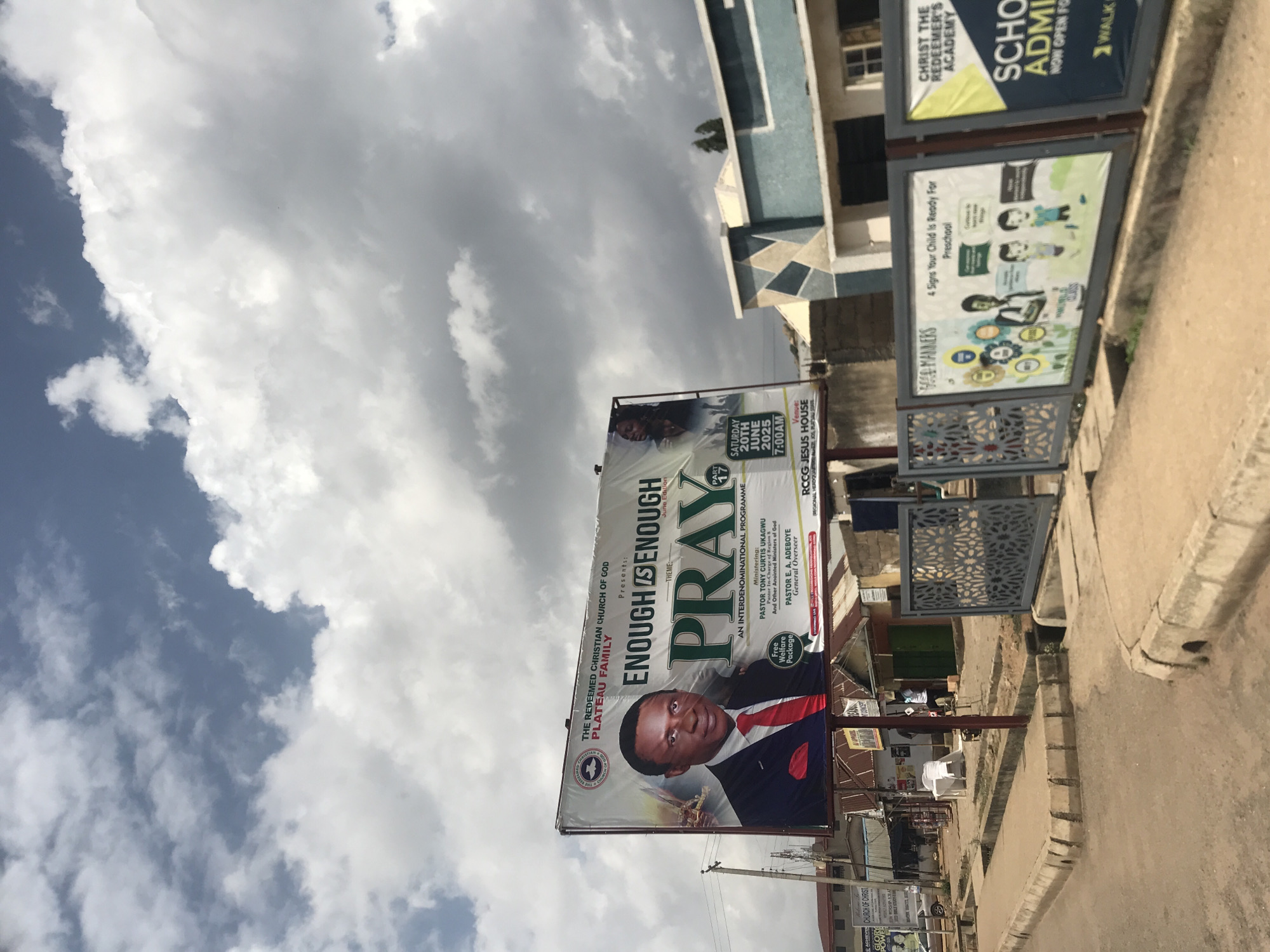 The image shows a street scene in New Stadium, Nigeria, with a prominent billboard advertising an event by The Redeemed Christian Church of God, Plateau Family. The billboard features a large portrait of Pastor Tony Curtis Ukagwu and text promoting an interdenominational program titled "Enough is Enough: Pray". The event is scheduled for Saturday, June 20th, 2025, at 7:00 AM, at RCCG Jesus House. The billboard also mentions a "Free Welfare Package" and Pastor E.A. Adeboye, General Overseer. To the right of the main billboard, there are two smaller billboards. One advertises "Good Manners" with illustrations about preparing a child for preschool, and the other announces "Christ the Redeemer's Academy" and "School Admissions". The street is paved with concrete and lined with buildings, some of which appear to be shops. There are also some parked cars and a few people visible in the background. The sky is filled with dramatic clouds.