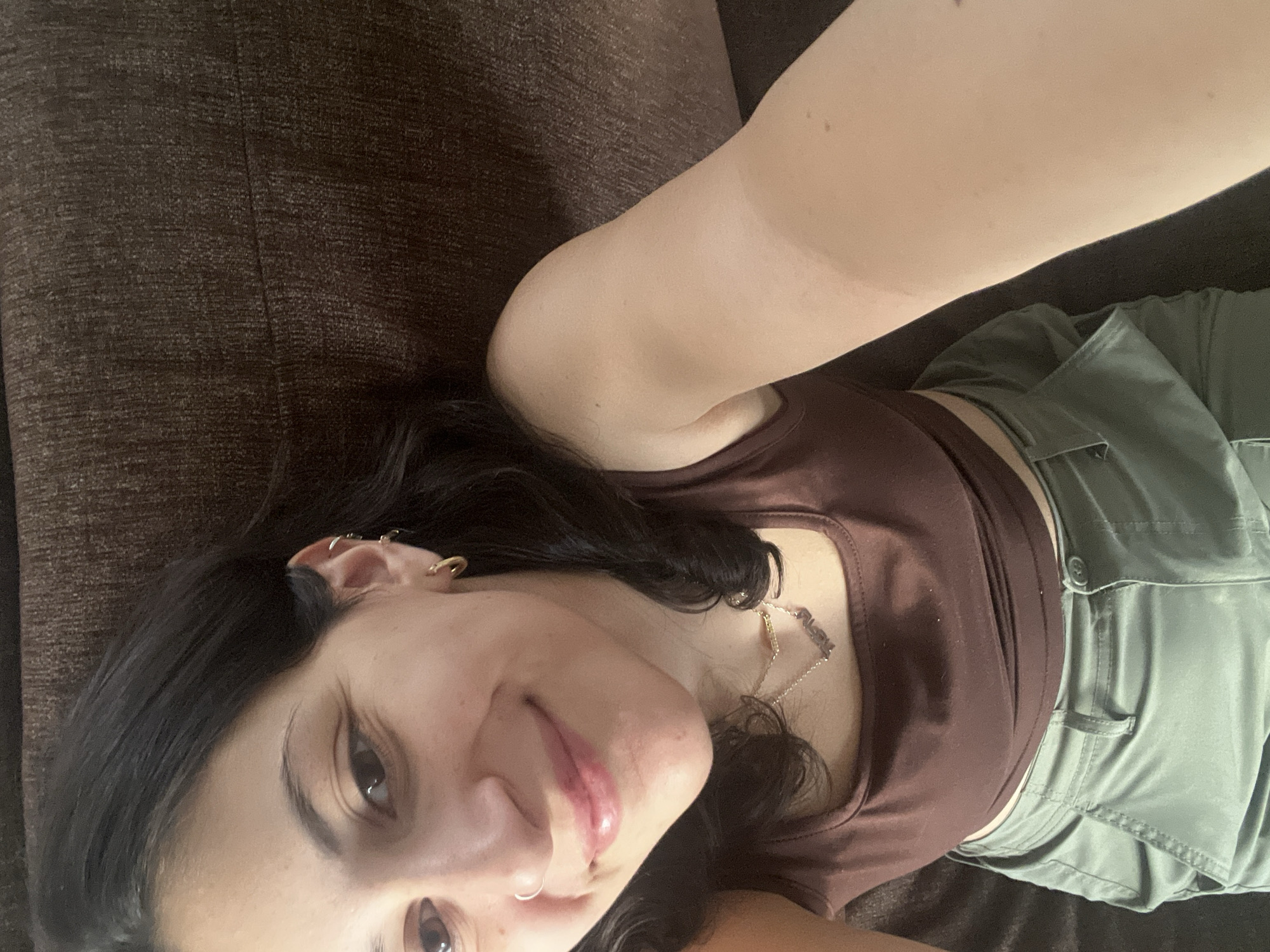 A woman with dark hair is lying on a dark brown couch. She is wearing a brown tank top and green shorts. She has earrings in her ear and a necklace around her neck. The location is unknown, but it is likely in Mexico City, Mexico.